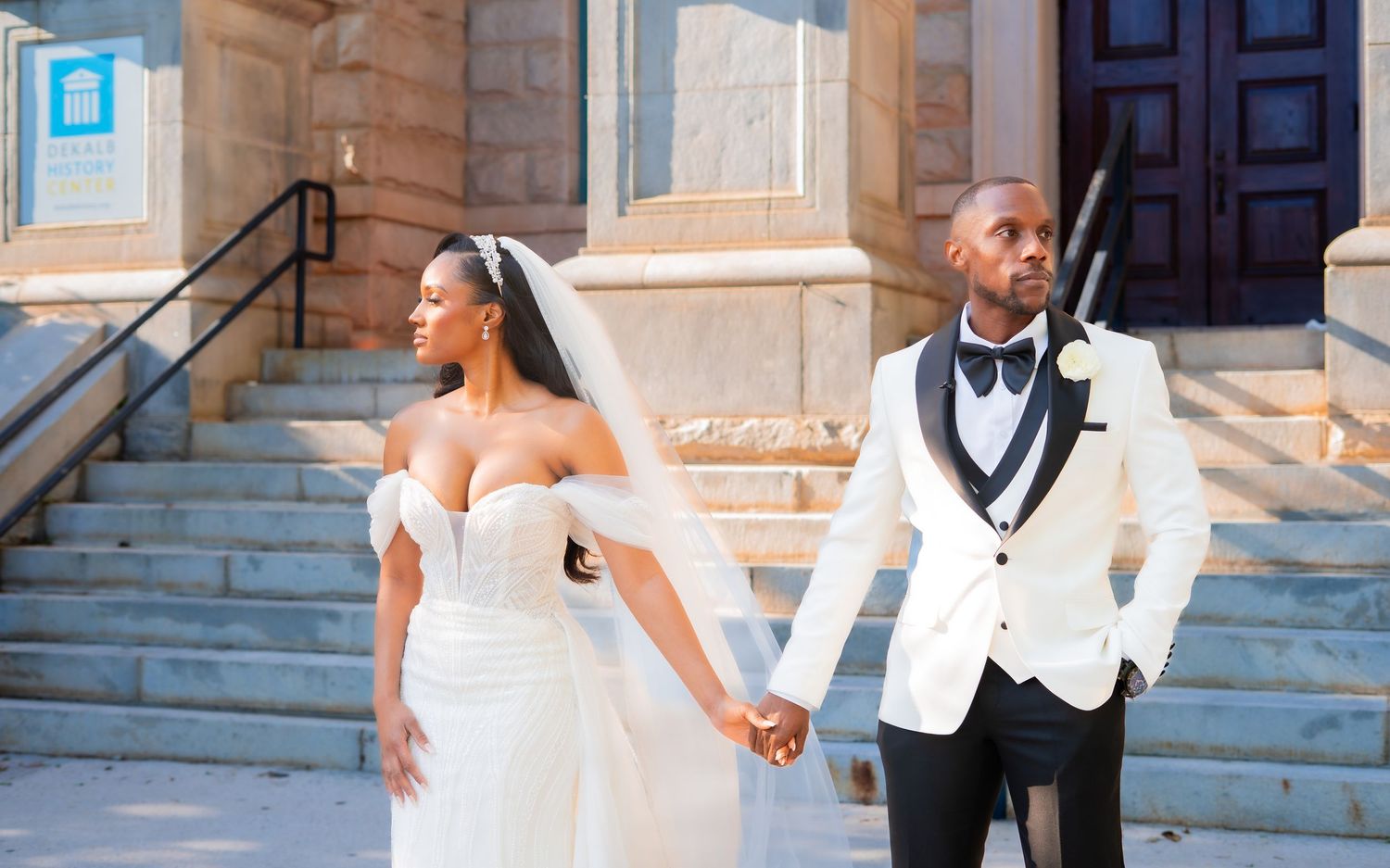 A happy wedding couple poses together on stone steps with white dress and black tuxedo in an elegant outdoor setting.