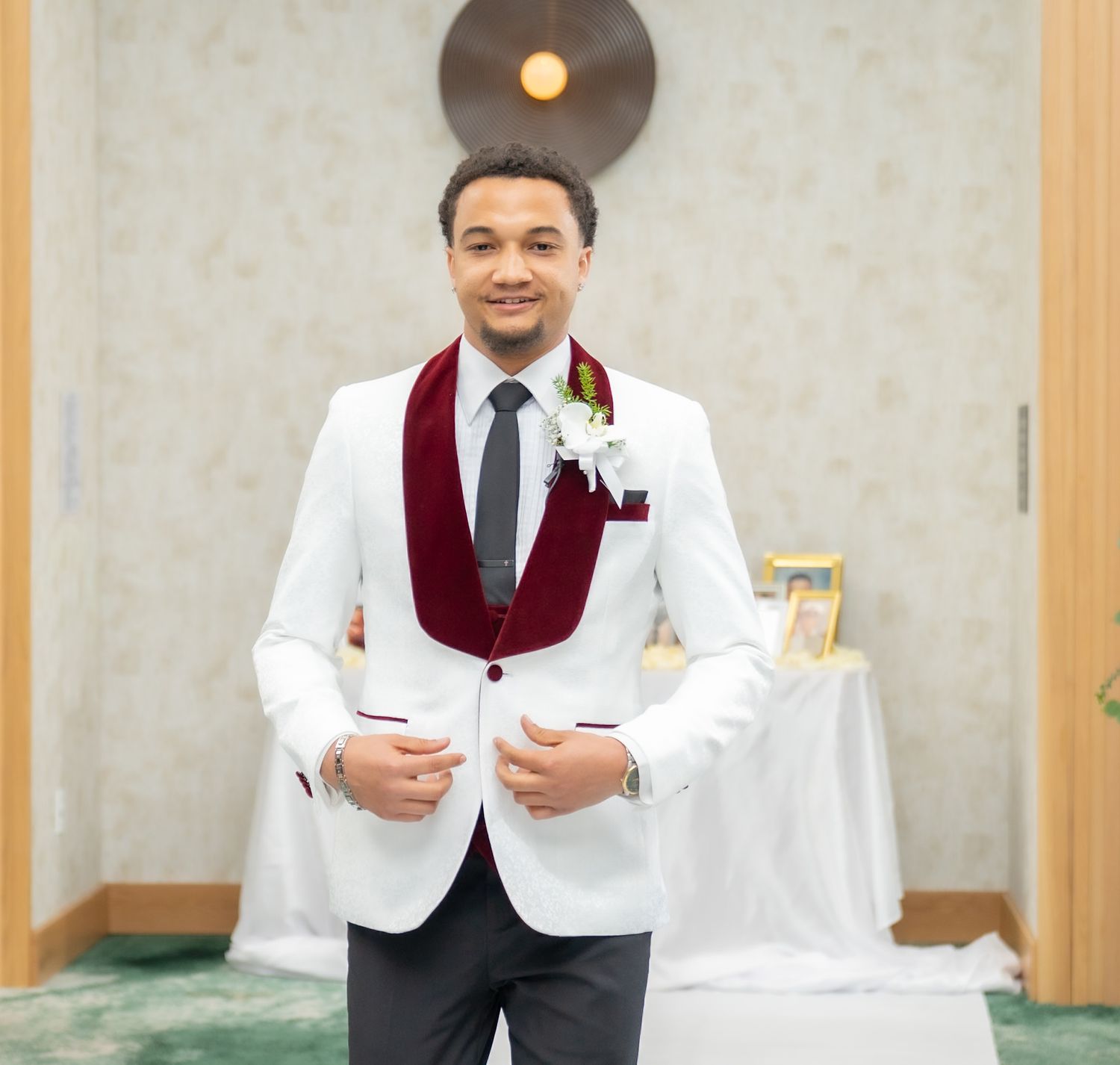 Man in white tuxedo jacket with burgundy velvet lapels and floral boutonniere posing at a formal event.