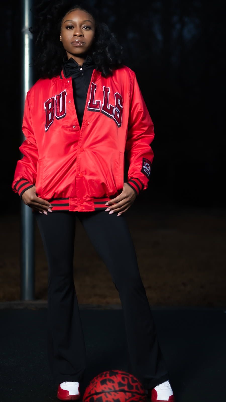 Someone wearing a bright red Chicago Bulls satin bomber jacket poses against a dark background in black pants and sneakers.