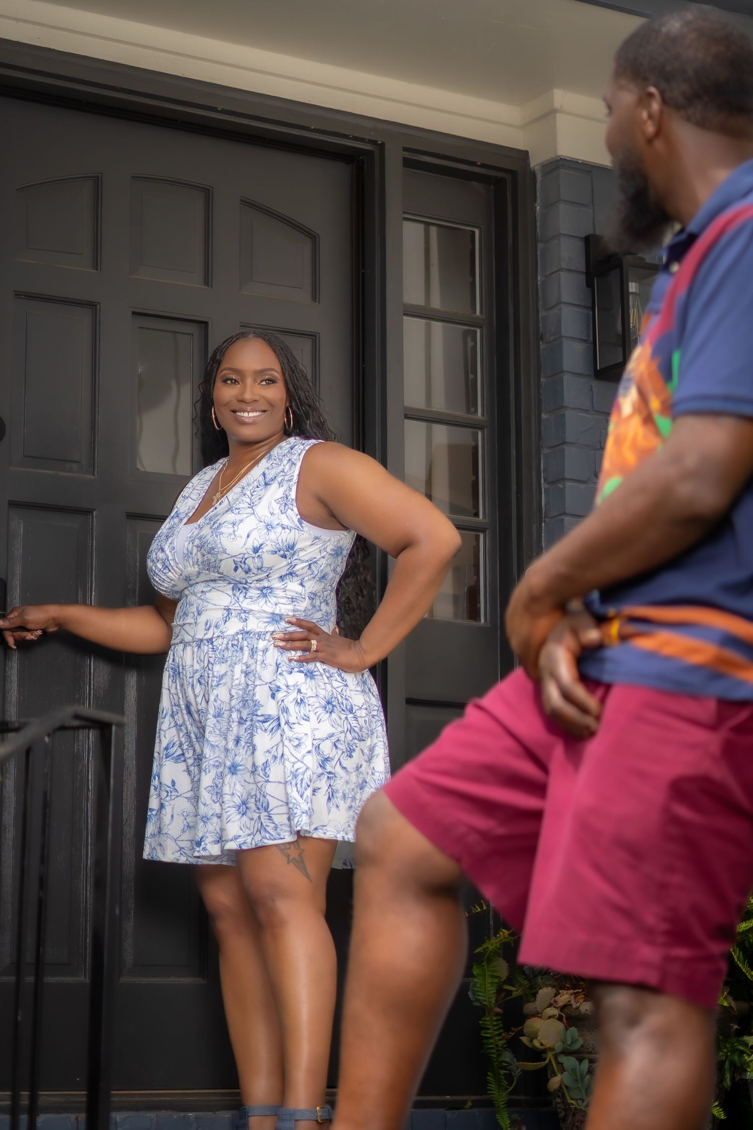 A smiling woman in a blue floral dress poses confidently at a dark front door while a man in red shorts looks on.