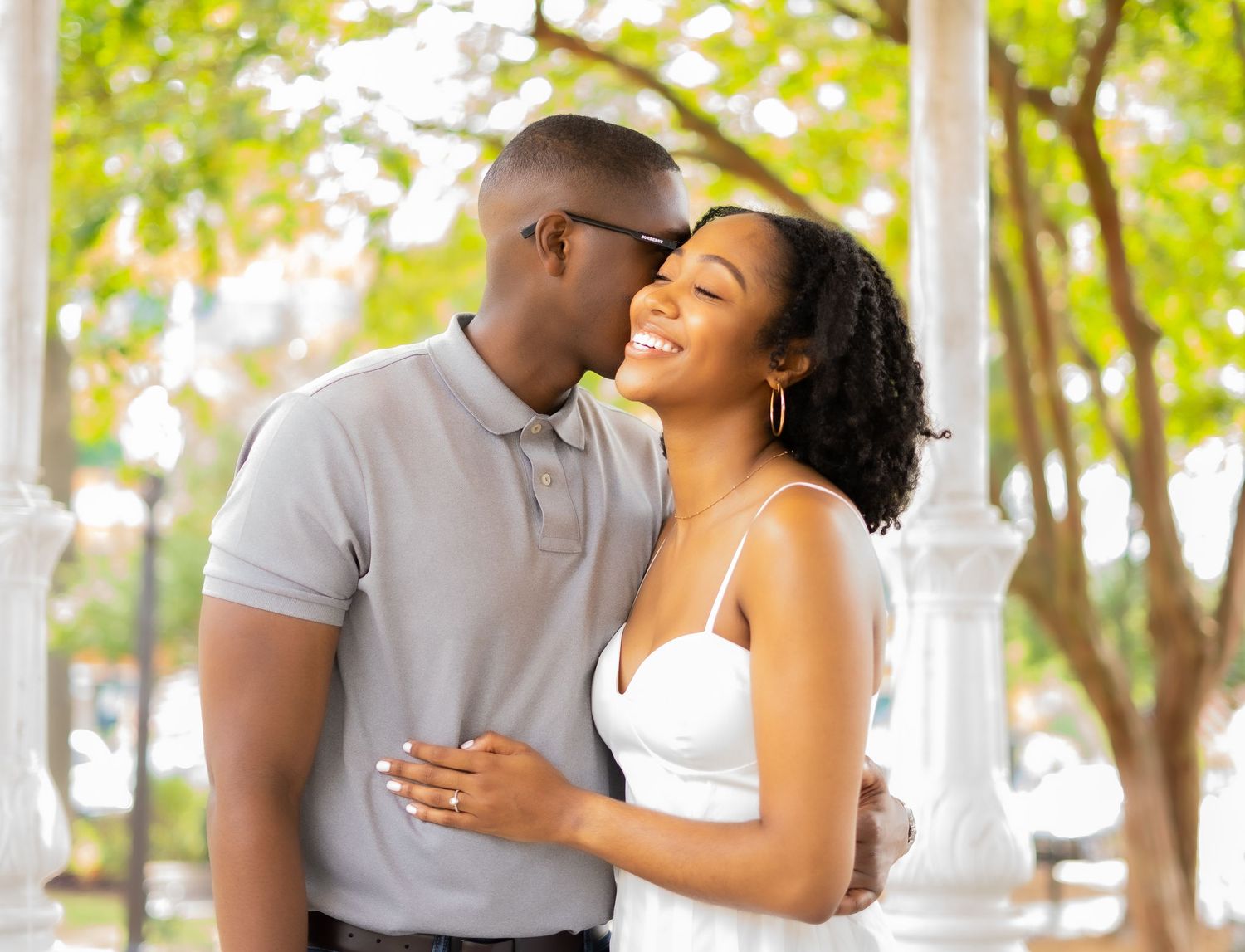 A loving couple shares a tender moment outdoors on a sunny day surrounded by trees in a romantic setting.