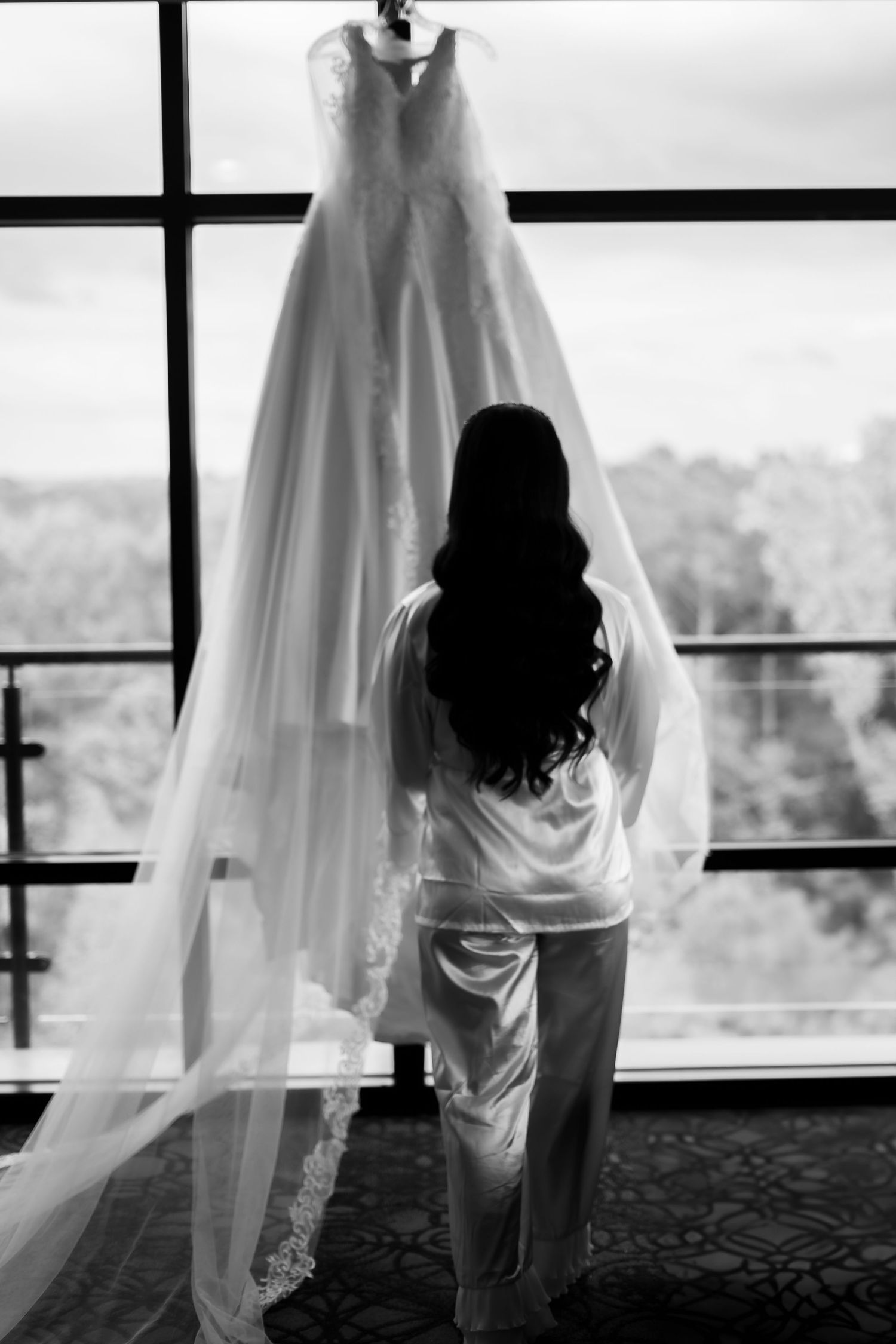 Silhouette of bride in robe looking at wedding dress displayed by window.