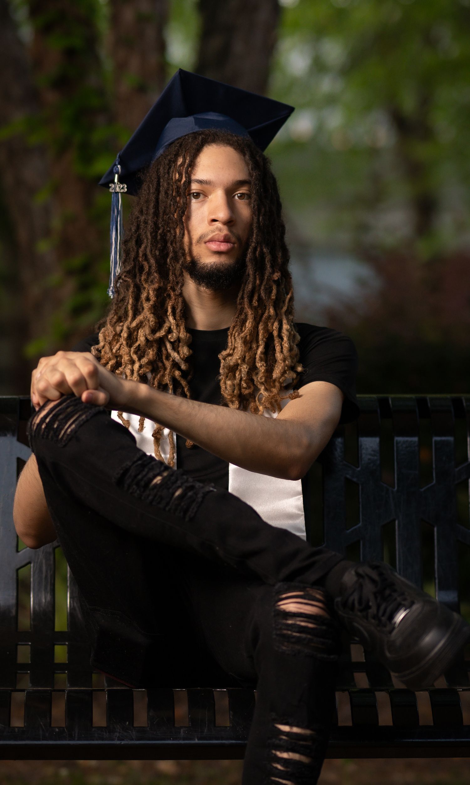 A graduate in a black cap and gown sits on a park bench wearing ripped jeans and casual attire.