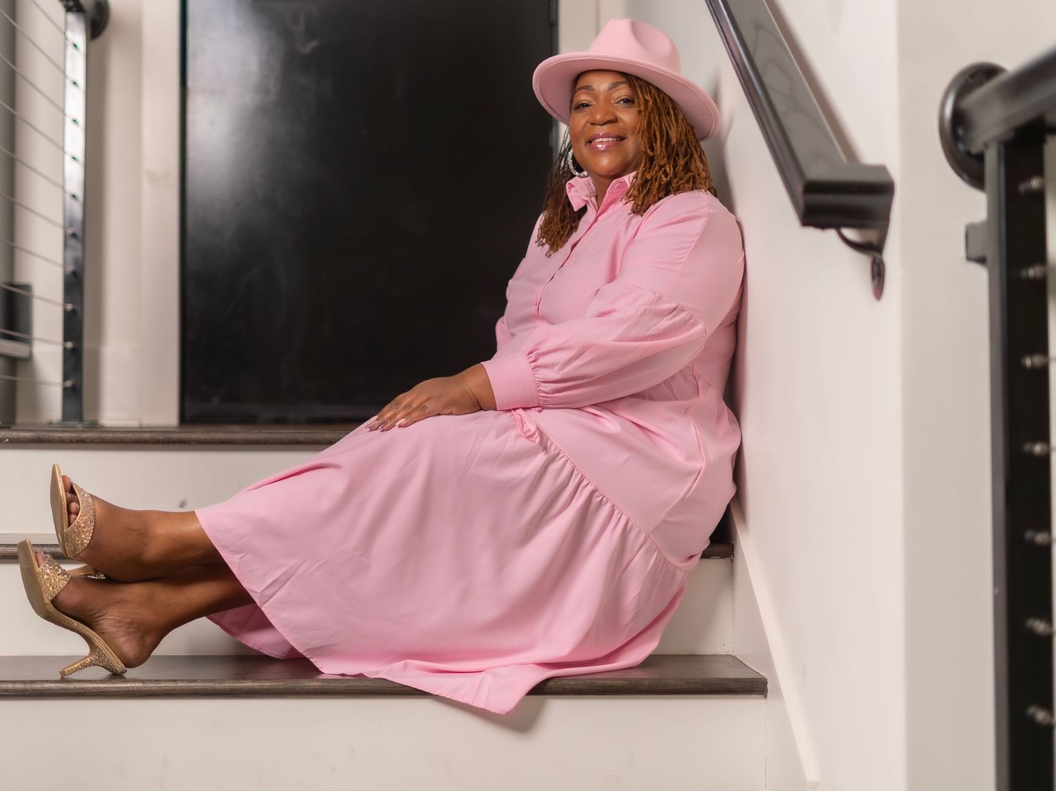 Someone in a pink western-style dress and cowboy hat sits casually on white stairs with brown heeled boots.