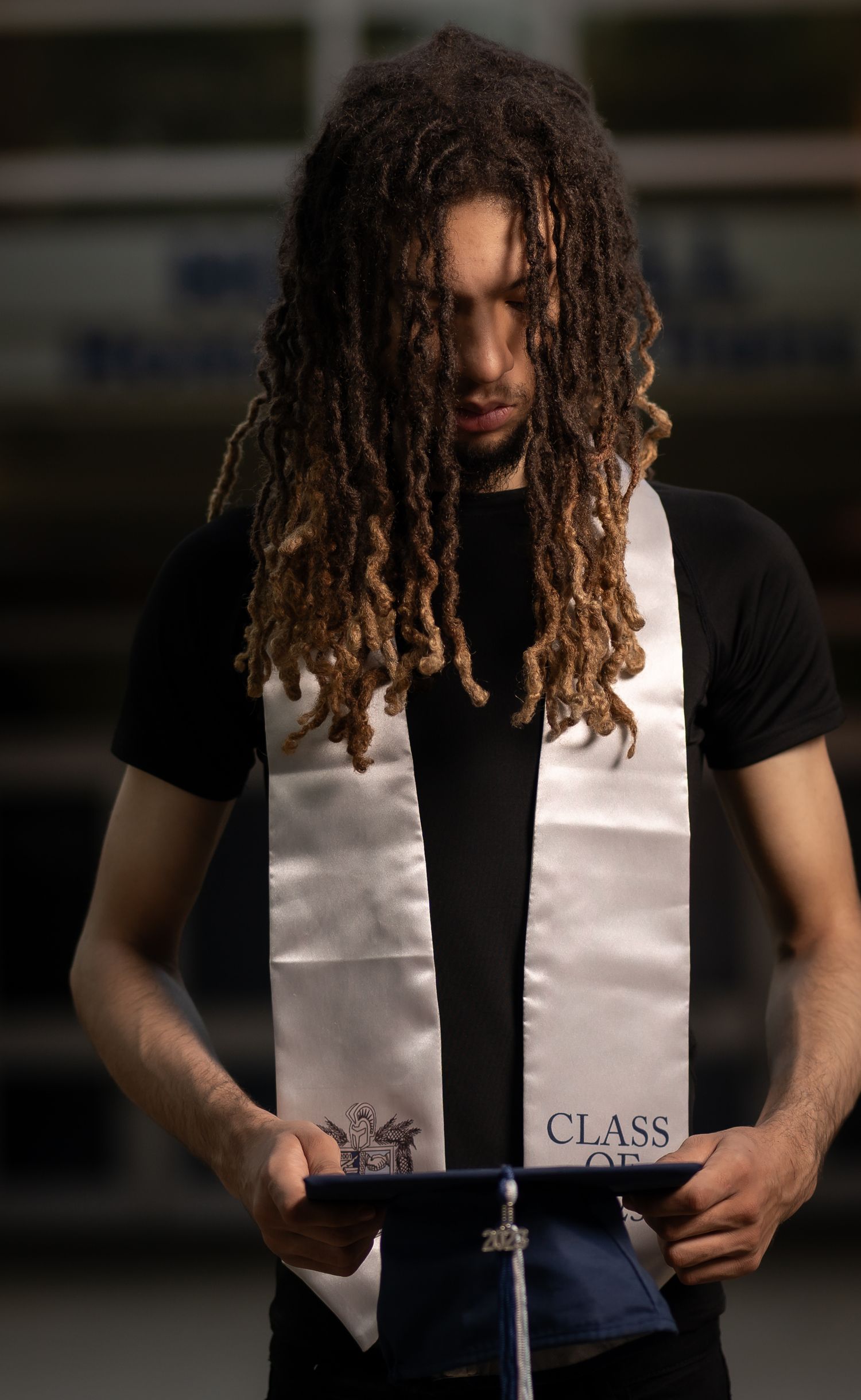 Person with long curly dreadlocks wearing a black t-shirt and white vest holding a book against dark background.