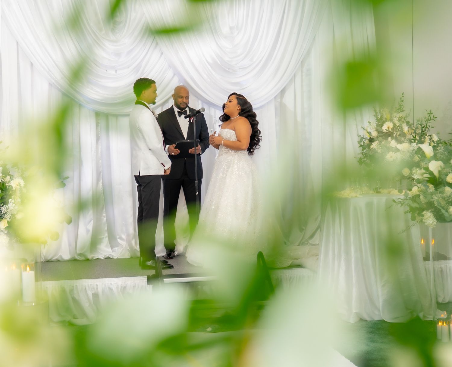 Bride and groom exchange vows before officiant at elegant white-draped wedding ceremony altar with floral arrangements.
