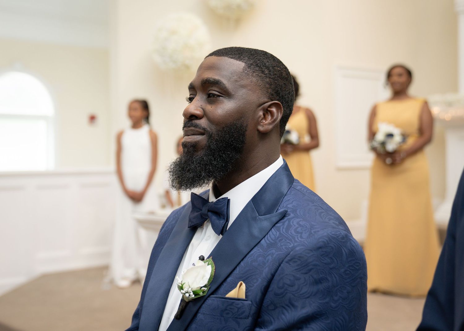 A joyful groom in a navy suit and bow tie smiles warmly during a wedding ceremony.