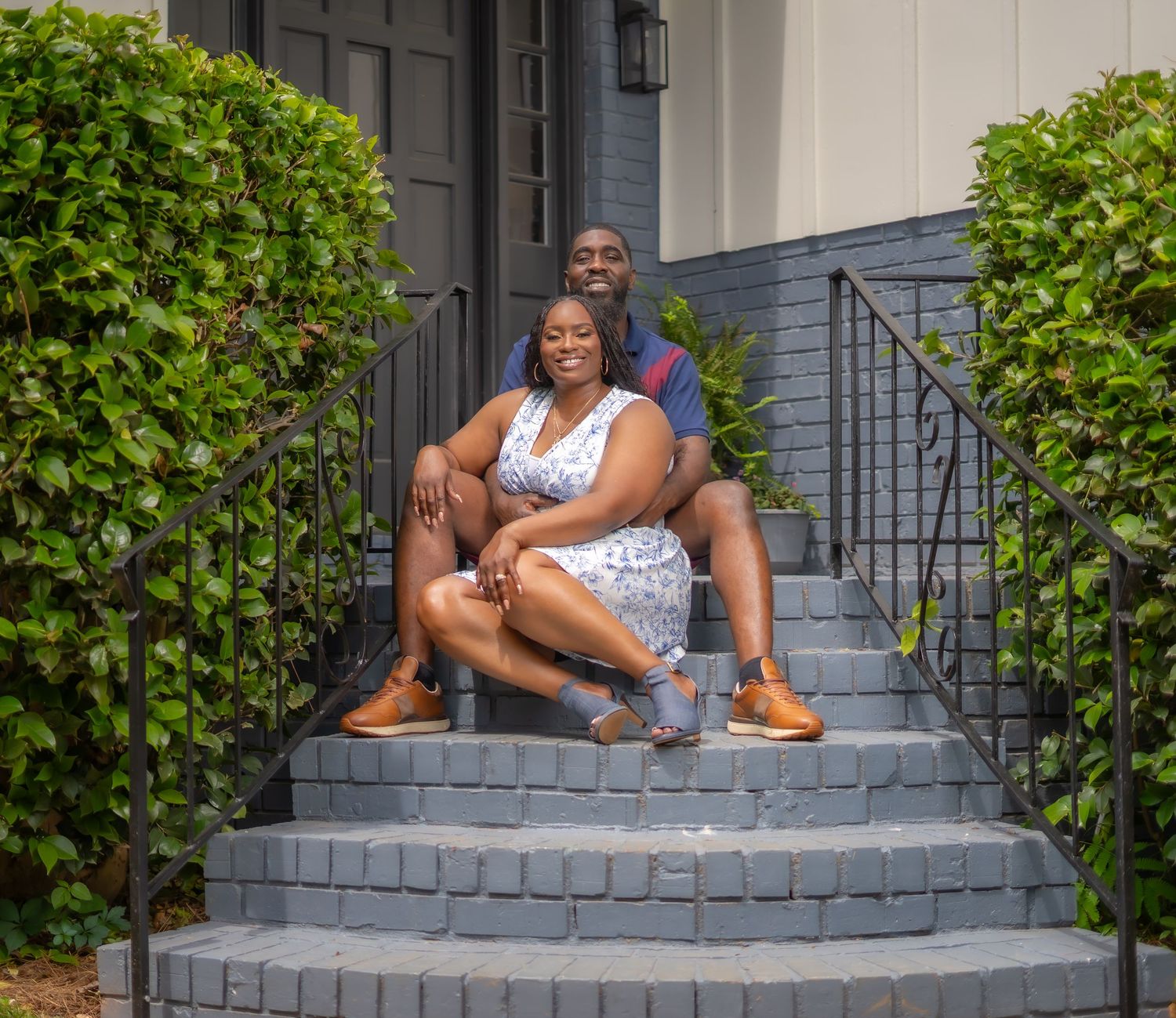 Couple sitting together on gray brick front steps of a home, surrounded by green bushes on a sunny day.
