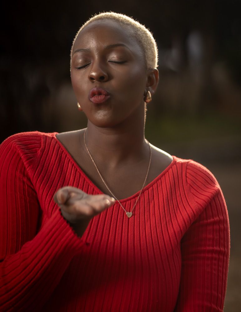 Woman in red ribbed sweater with short blonde hair blowing a kiss, wearing a gold heart necklace outdoors.