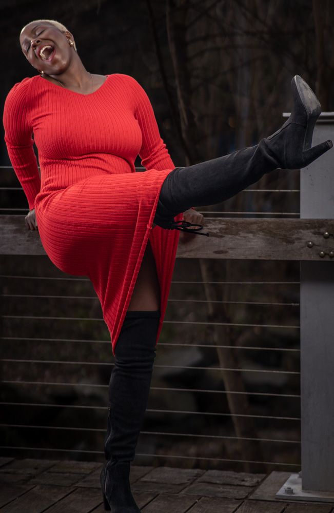 Woman in red ribbed dress and black thigh-high boots posing confidently on an outdoor bridge railing.