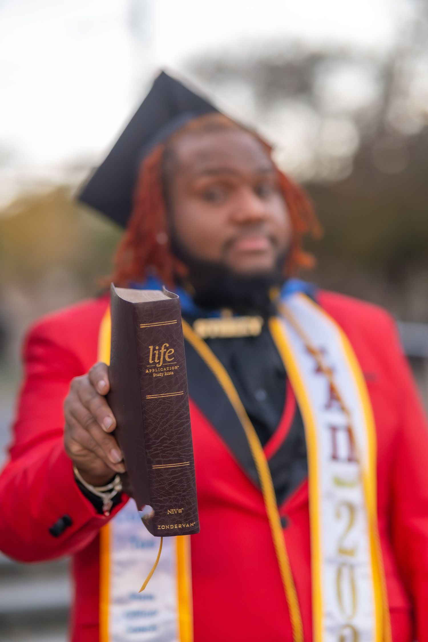 A person in red and gold graduation regalia holds up a small book while standing outdoors.