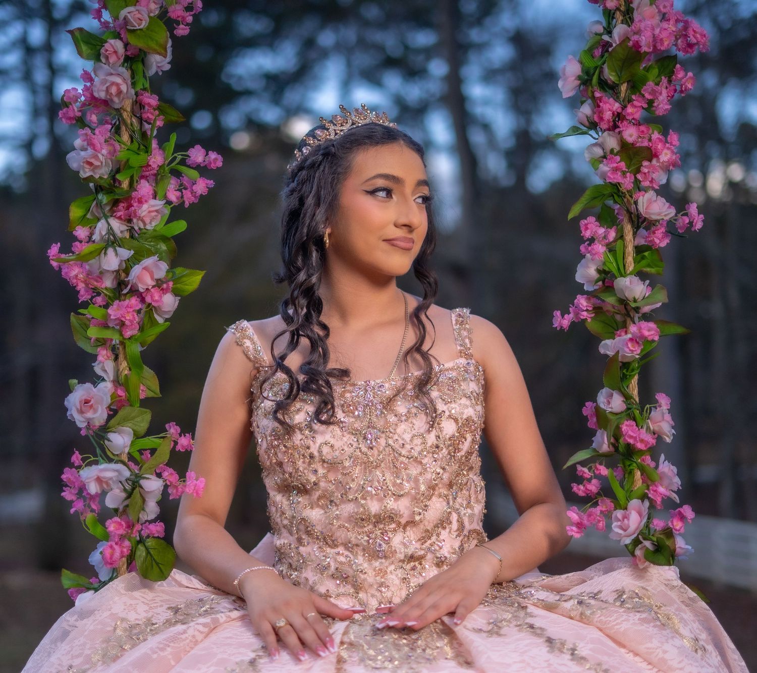 A person in a sparkling rose gold quinceanera gown poses elegantly in an outdoor garden setting with pink flowers.