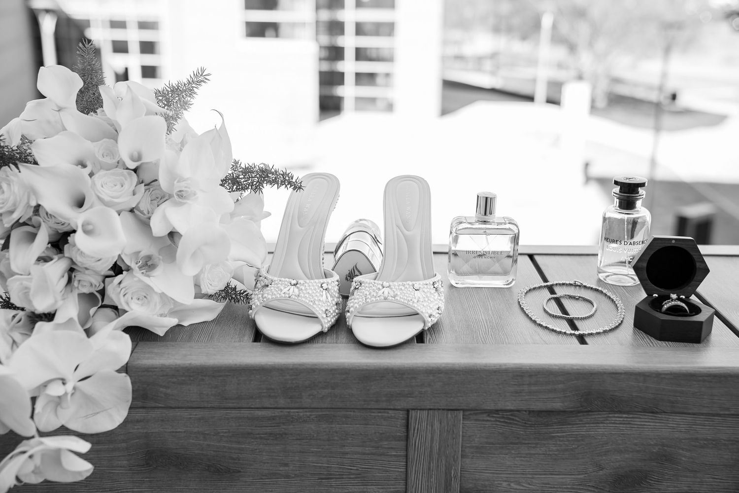 Black and white photo of bridal accessories including heels, perfume bottles, a ring box, and floral bouquet on a wooden dresser.