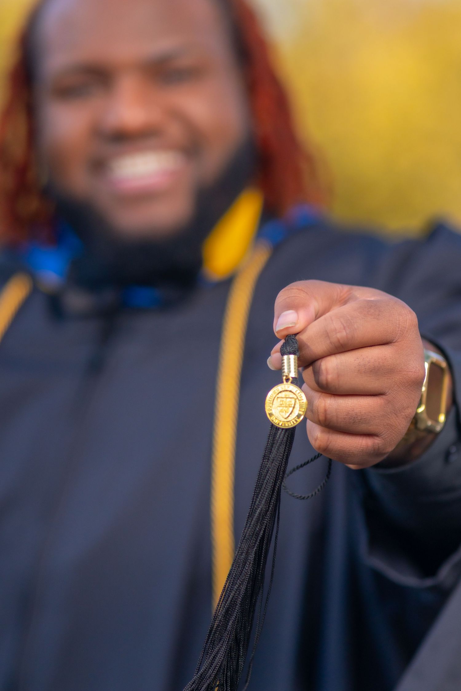 A graduate holding a black tassel with a gold medallion during commencement celebration in bright outdoor lighting.