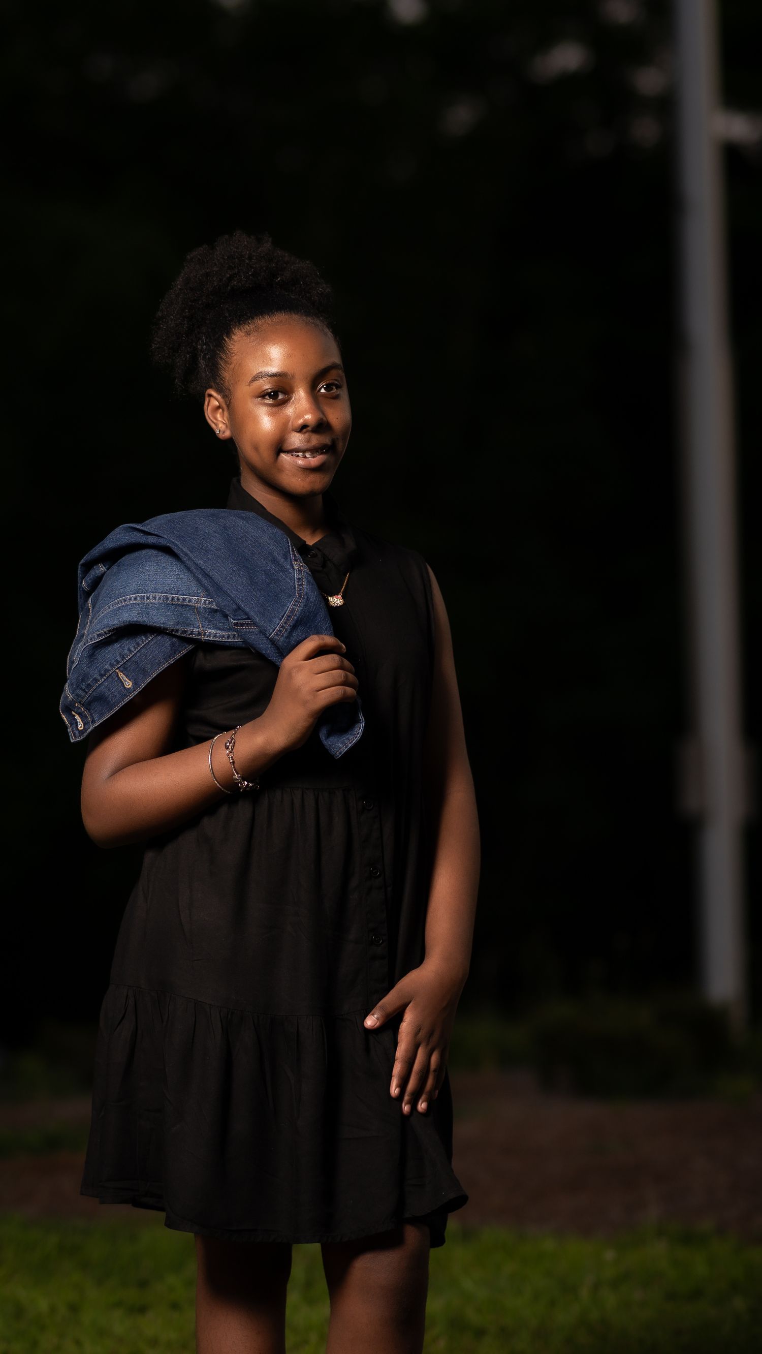 Young person in a black dress holds a denim jacket over their shoulder while posing outdoors at dusk.