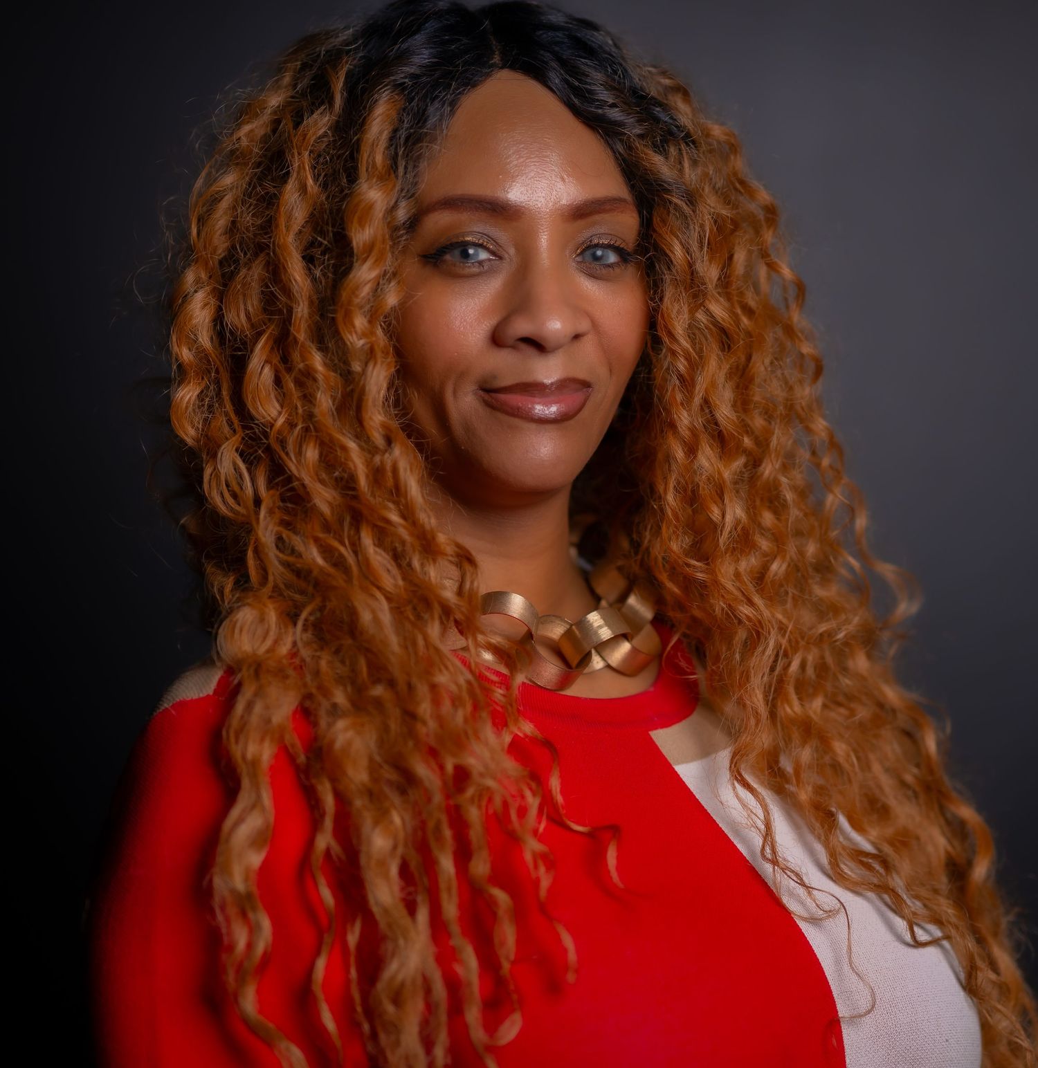 Person with long curly ombre hair wearing a bright red top poses against dark background.
