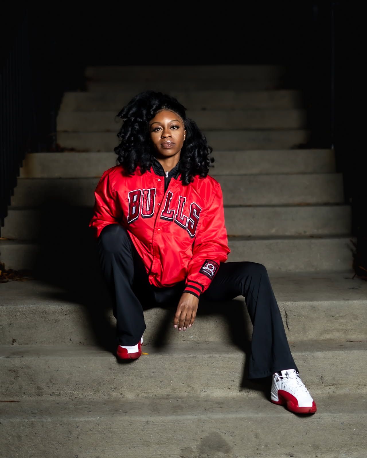 A person wearing a red Chicago Bulls satin jacket sits casually on concrete steps wearing white and red sneakers.