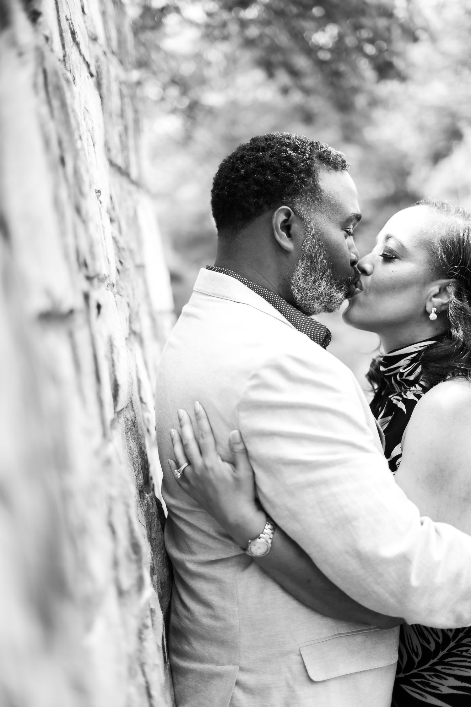 A black and white portrait captures a tender moment between a couple against a stone wall.