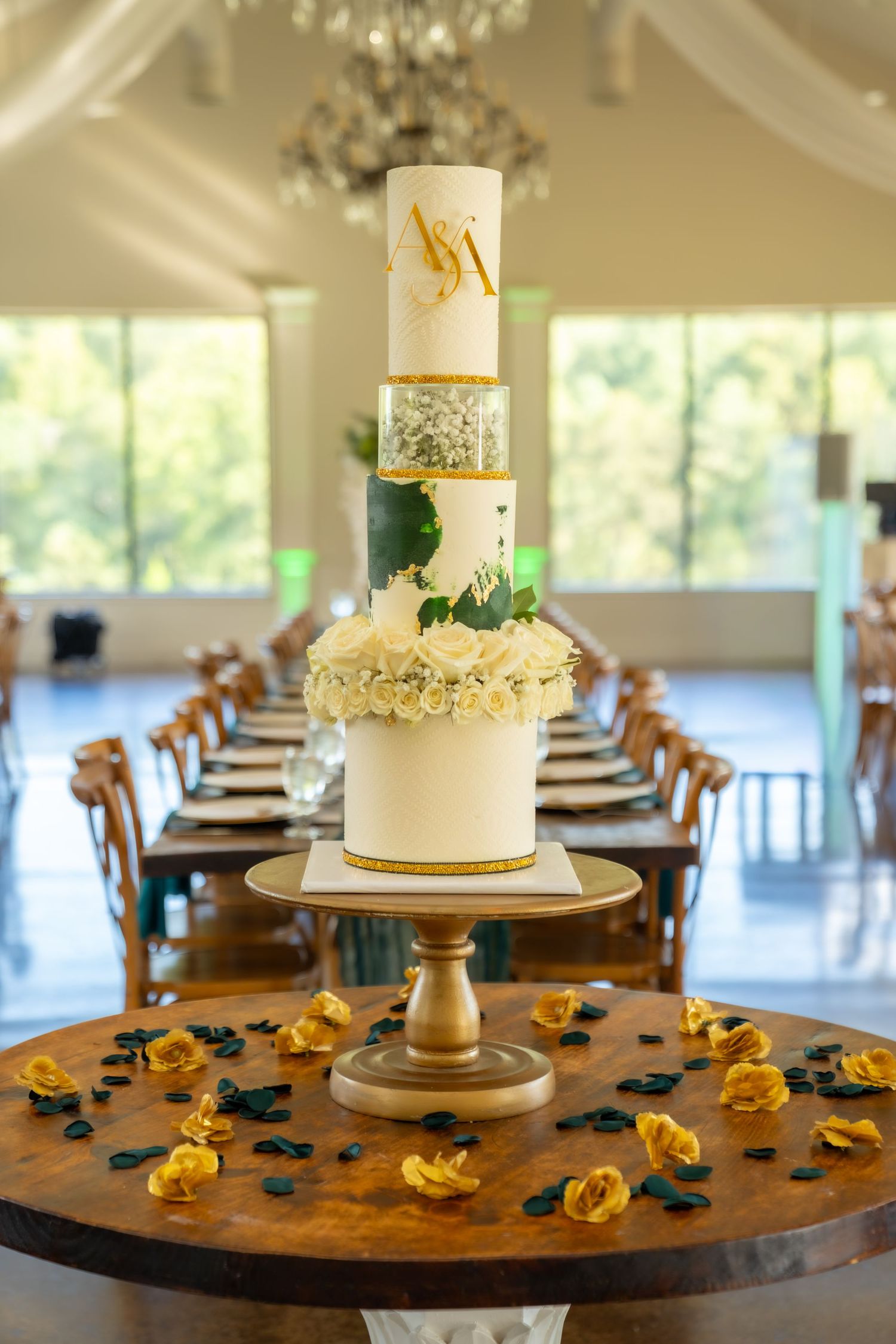 Elegant white wedding cake with gold accents and yellow rose petals scattered on rustic wooden table in reception venue.