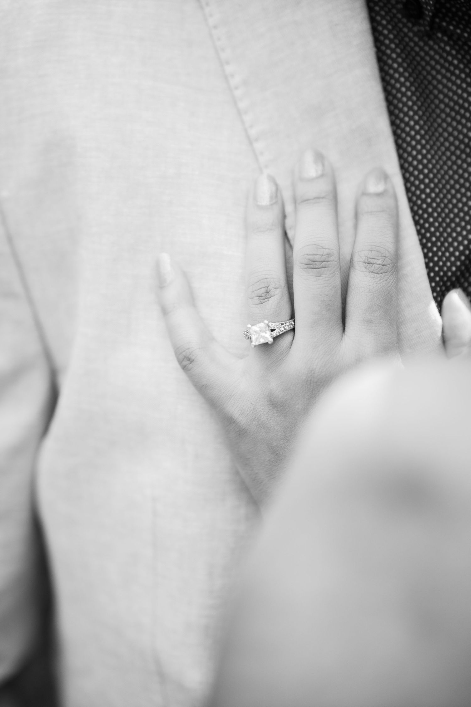 A black and white close-up photo of an engagement ring on a hand against a light background.