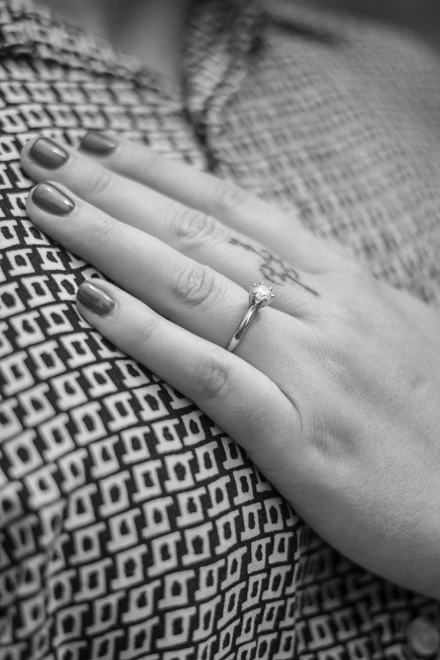 Close-up black and white photo showing a hand wearing an engagement ring resting on a patterned fabric.