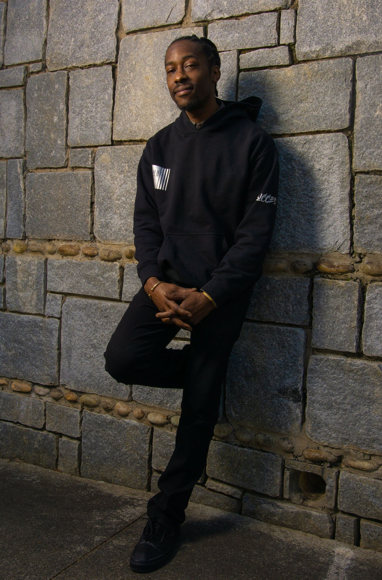 A person in an all-black outfit and hoodie leans against a stone wall while posing for a casual portrait outdoors.