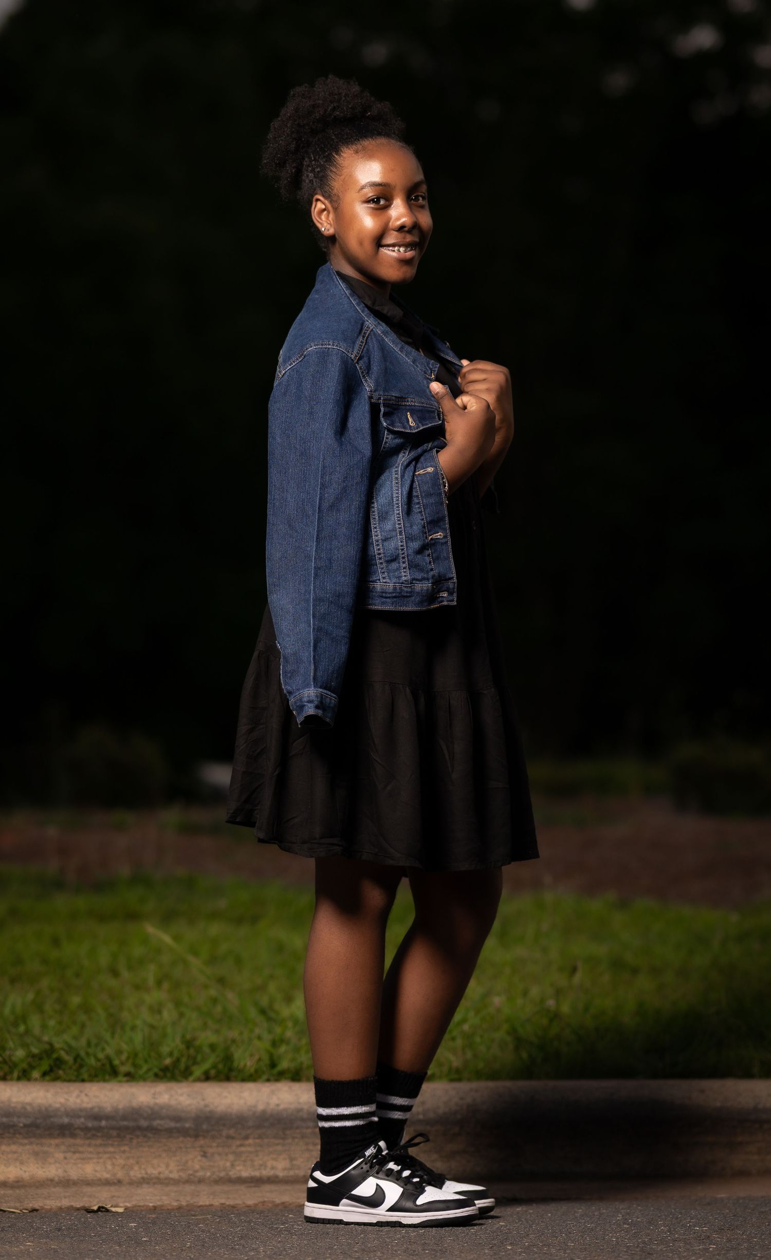 A stylish outfit featuring a denim jacket, black skirt, striped socks and sneakers against a dark outdoor background.