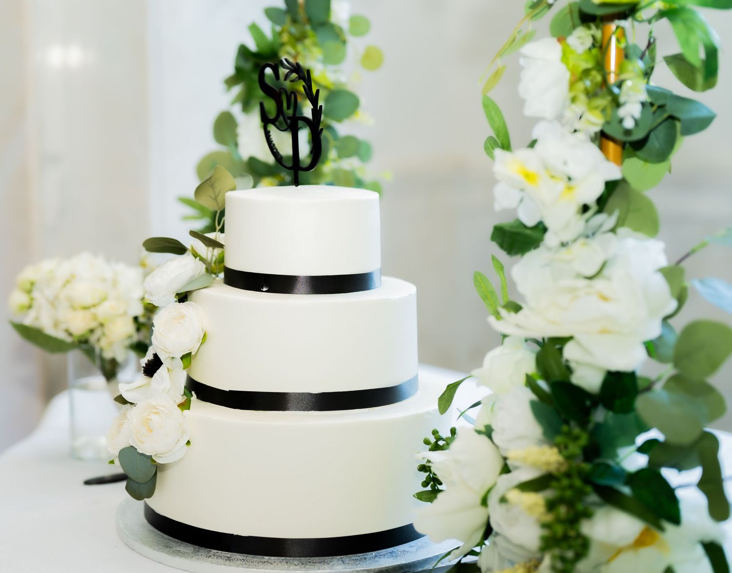 An elegant three-tier white wedding cake decorated with black ribbons and surrounded by white floral arrangements.