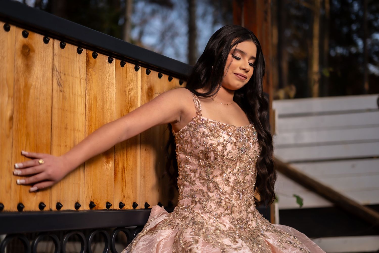 A person wearing a sparkly rose gold evening gown poses elegantly in front of a wooden fence at sunset.