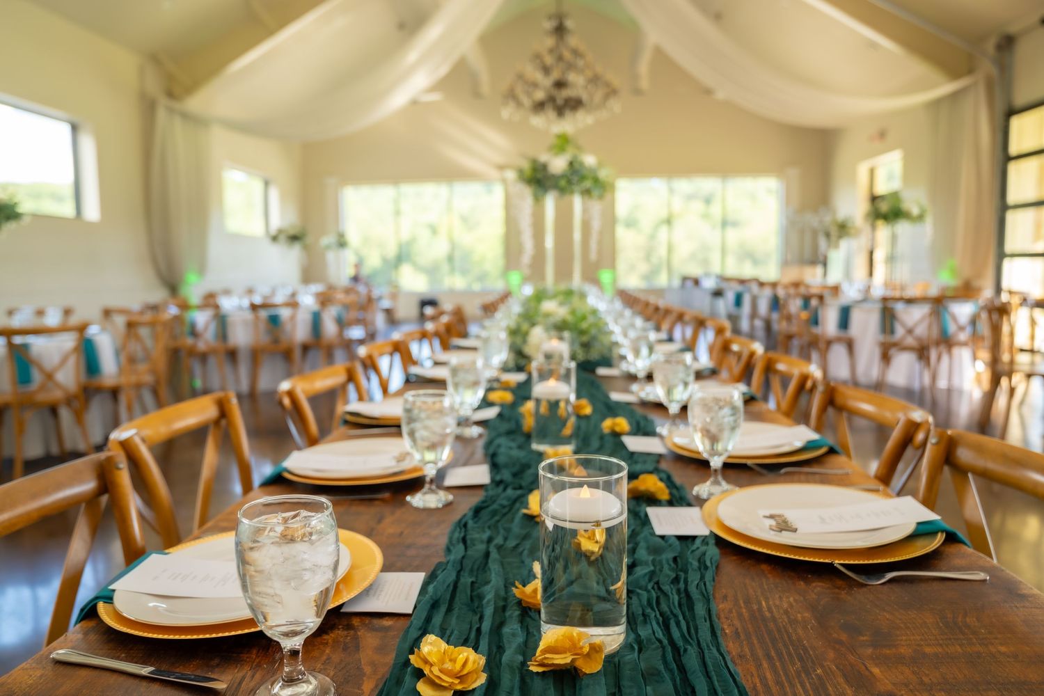 Long wooden dining tables set for an elegant wedding reception with teal runners and gold accents in a bright venue.