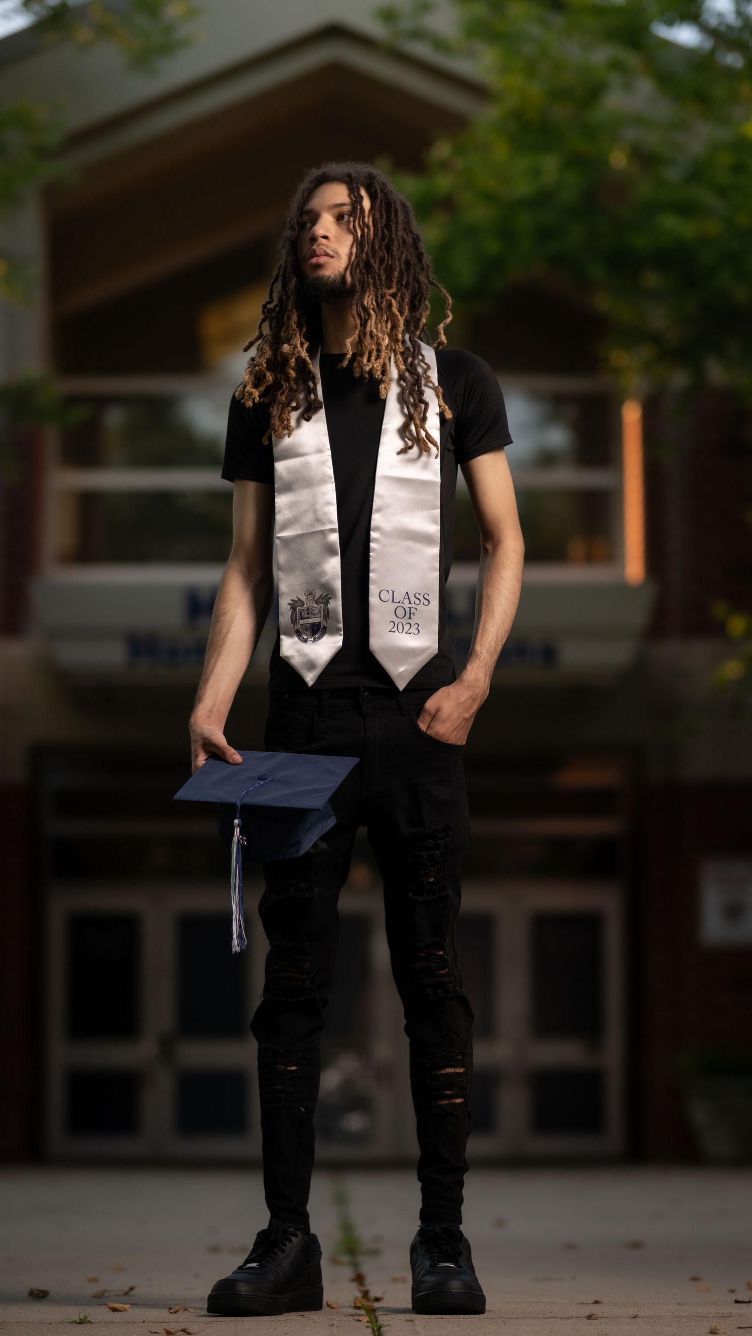 Graduate in black t-shirt and ripped jeans holds cap and wears graduation stole while standing outside building.