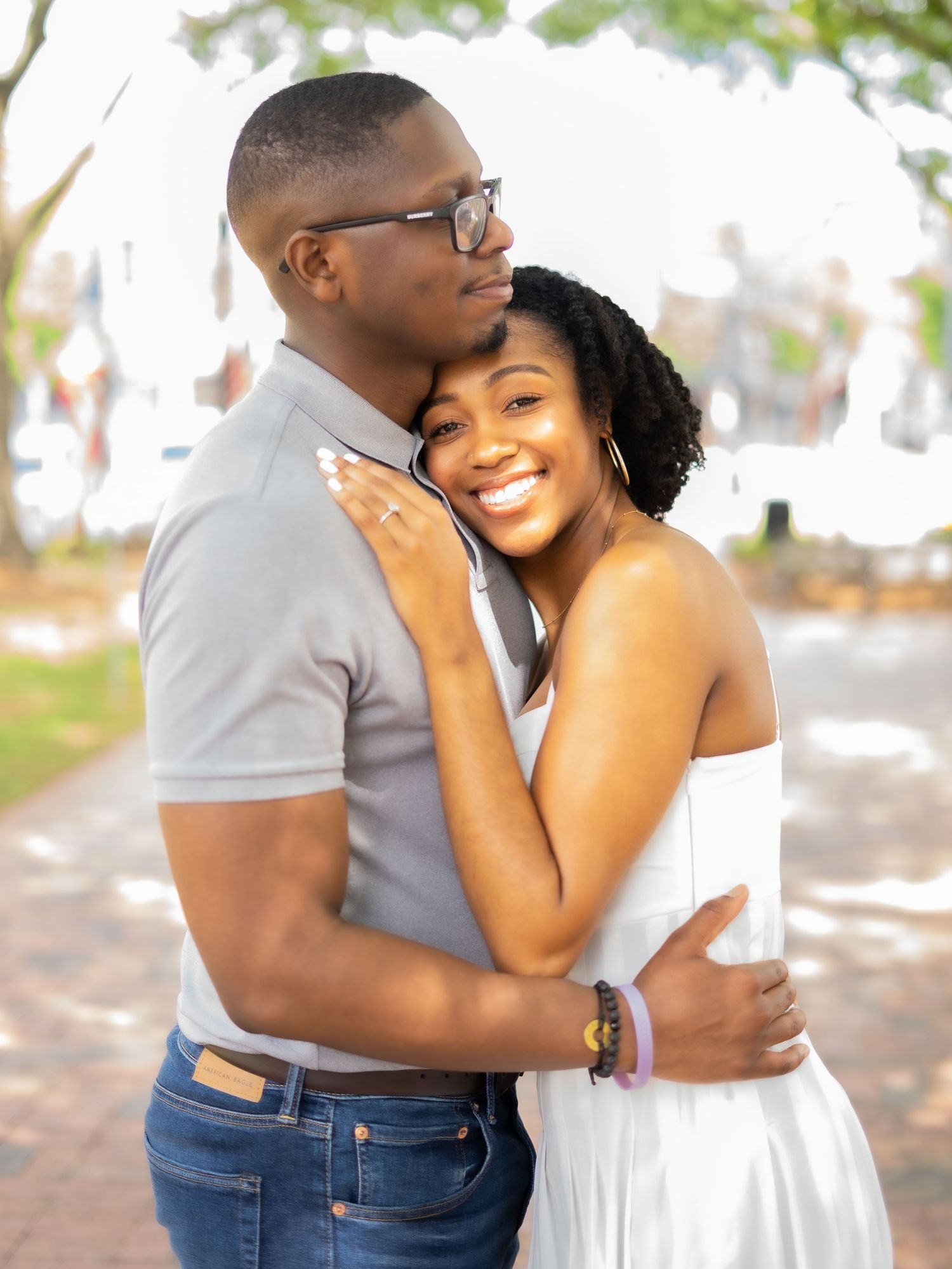 A romantic engagement photo of a couple embracing outdoors with sunlit trees in the background.