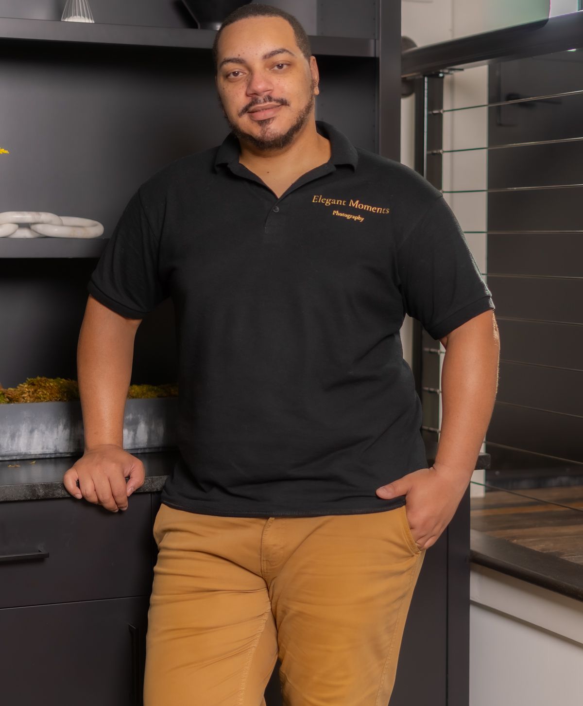 Restaurant worker in black uniform and khaki pants standing in modern dark commercial kitchen interior.