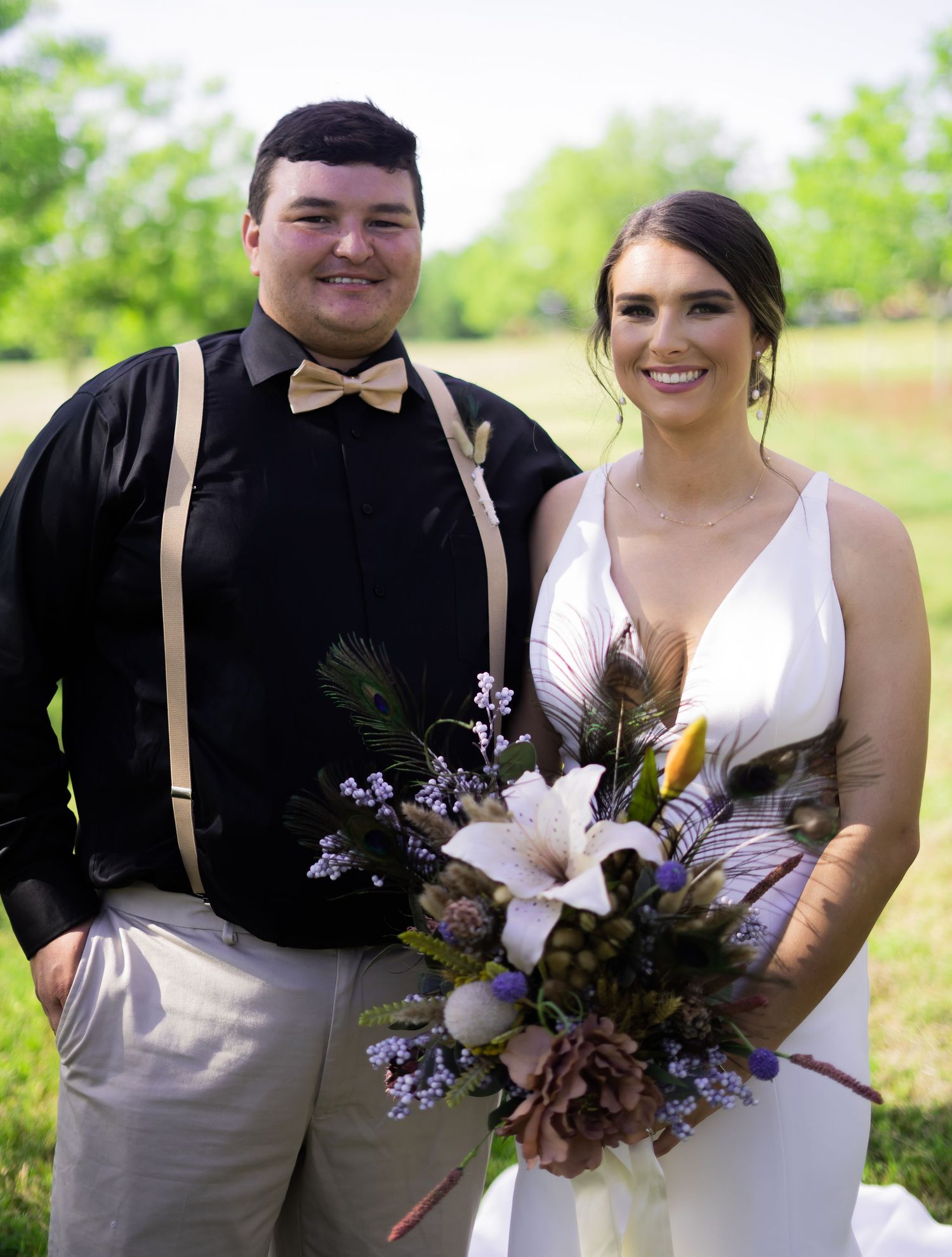 Outdoor wedding portrait with couple in casual attire featuring suspenders and a floral bouquet.