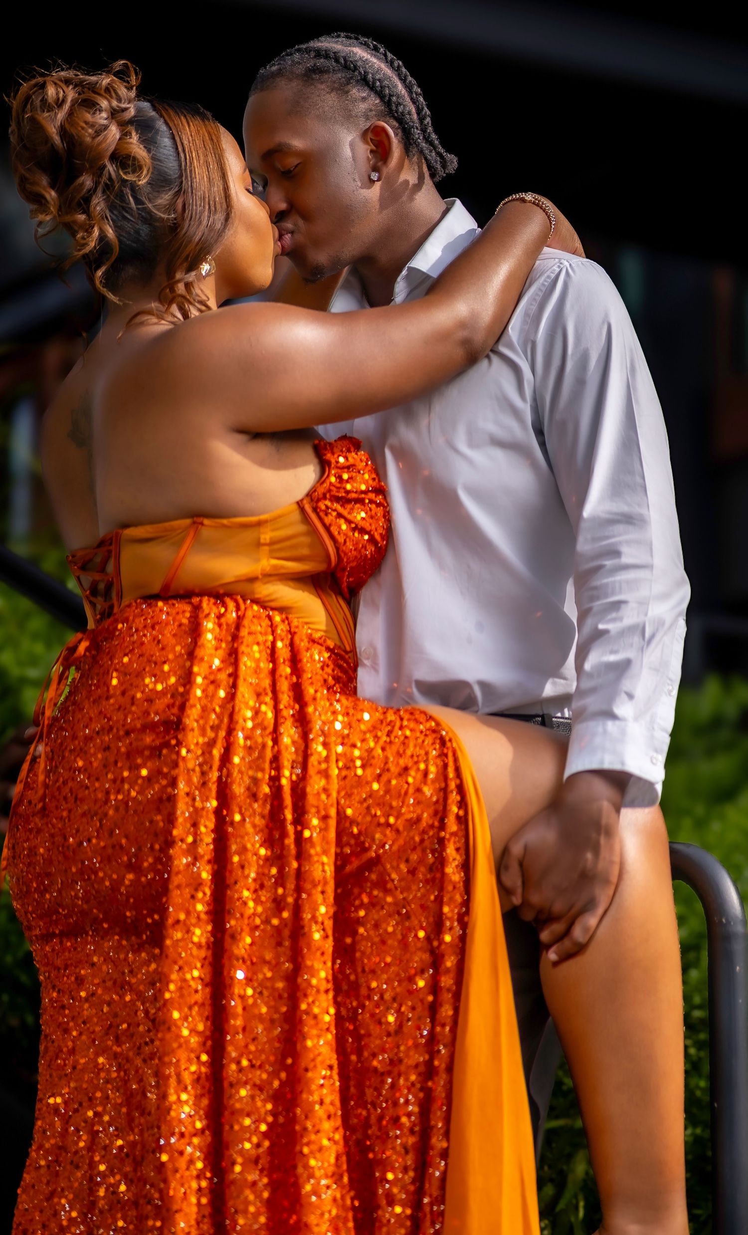 Couple sharing a romantic kiss, woman in orange sequin gown, man in white shirt, outdoor setting.