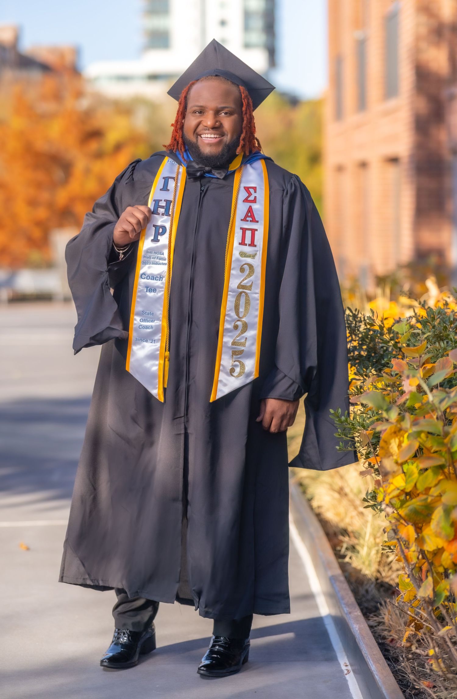 Graduate in black cap and gown with gold sash poses for graduation photos on campus during autumn season.