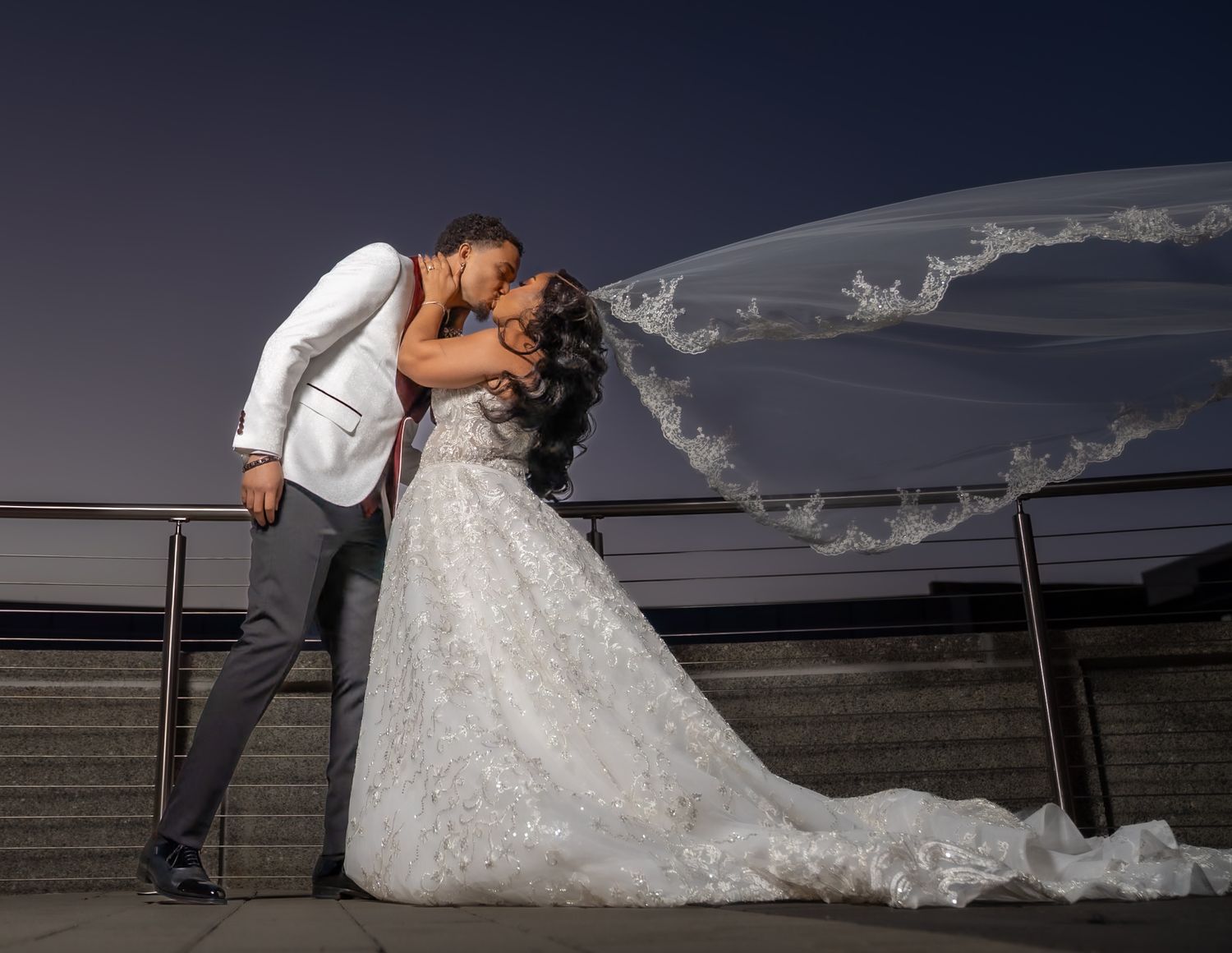 Bride and groom share a romantic kiss outdoors at night, with her lace wedding gown train and flowing veil billowing in the wind.