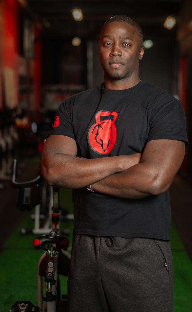 Athletic trainer in black t-shirt with red logo stands confidently with crossed arms in gym setting.