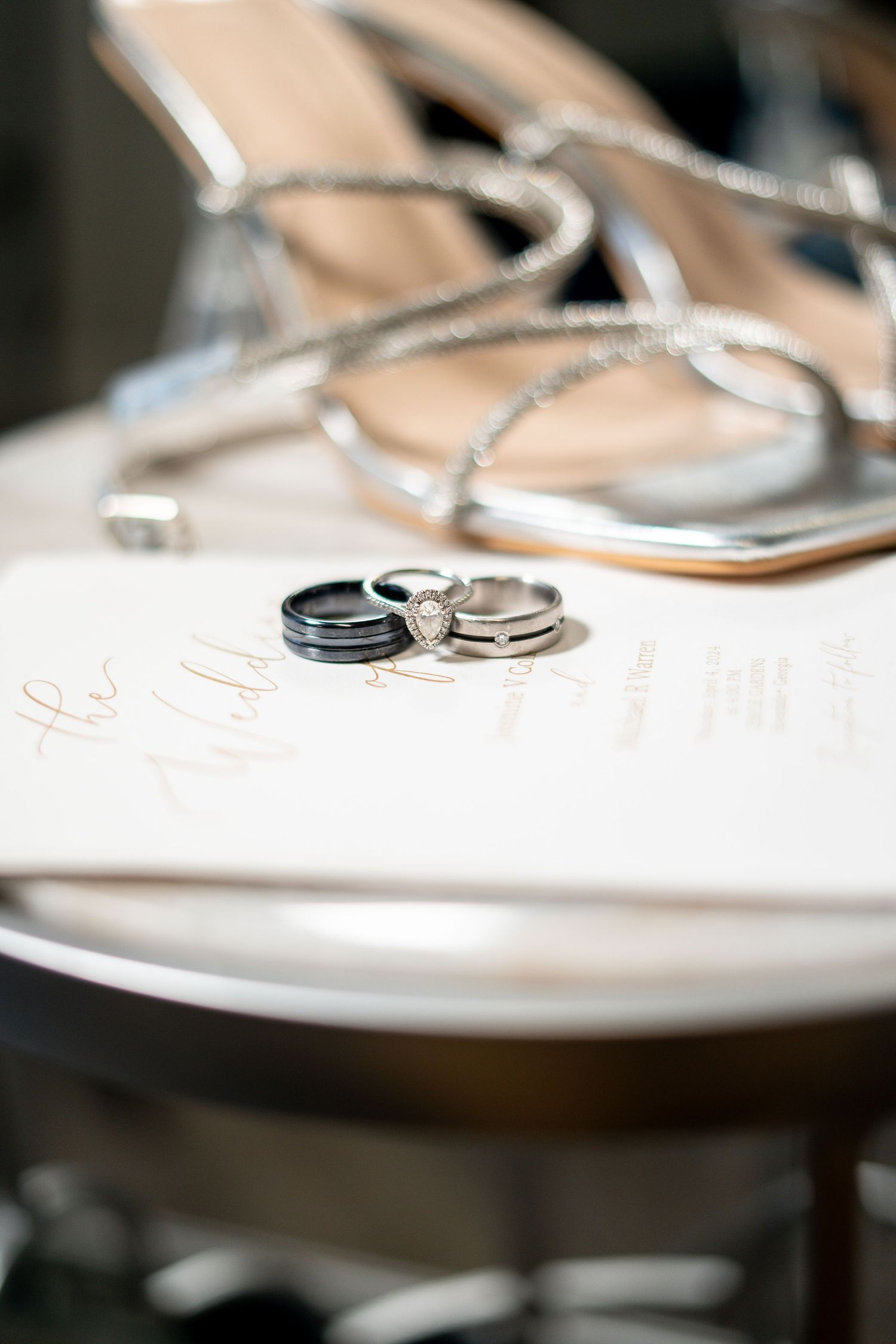 Wedding rings and silver sandals arranged on white surface in soft focus.