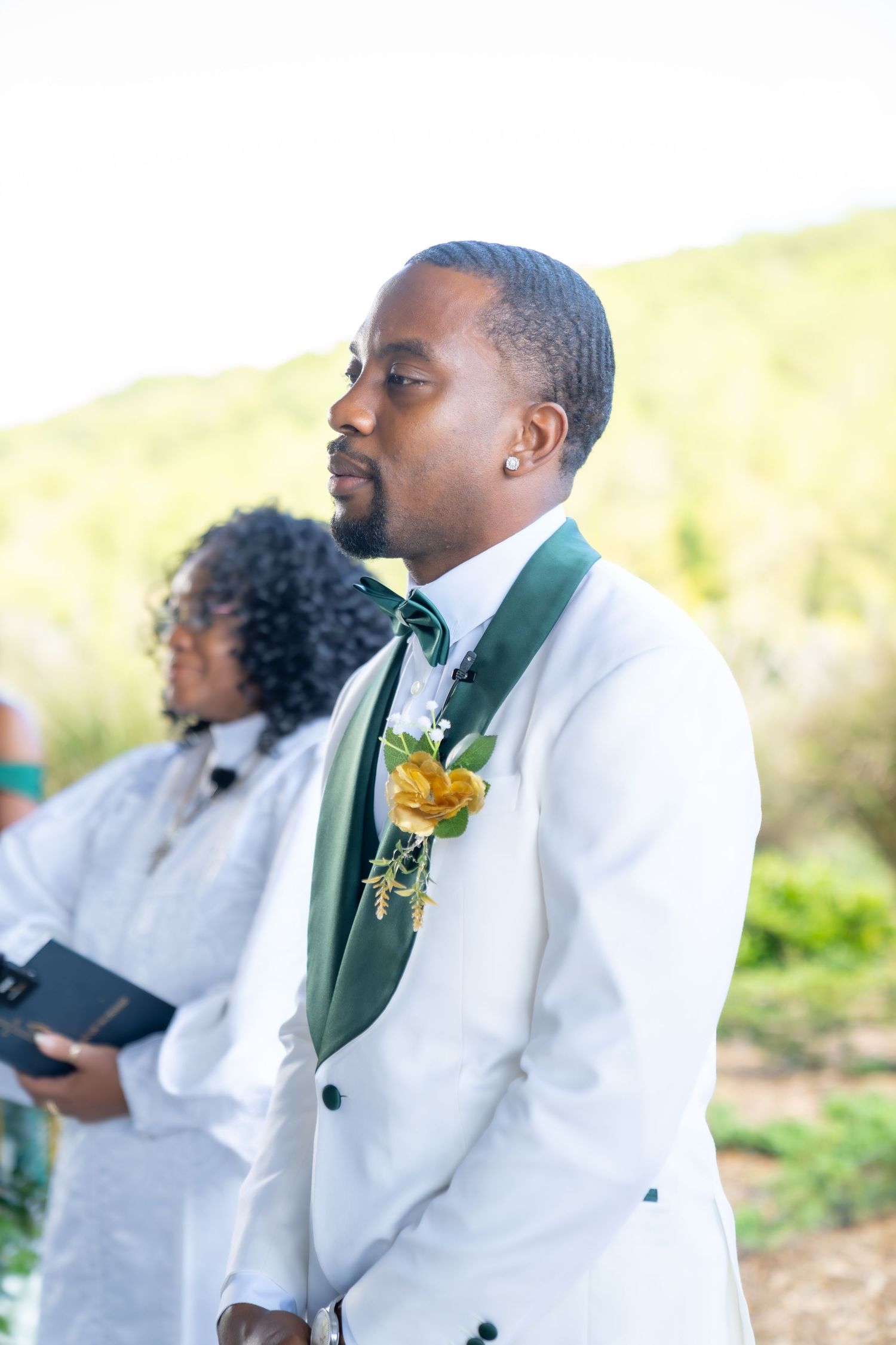 A groom in a white tuxedo with green lapels and yellow boutonniere smiles during an outdoor wedding ceremony.