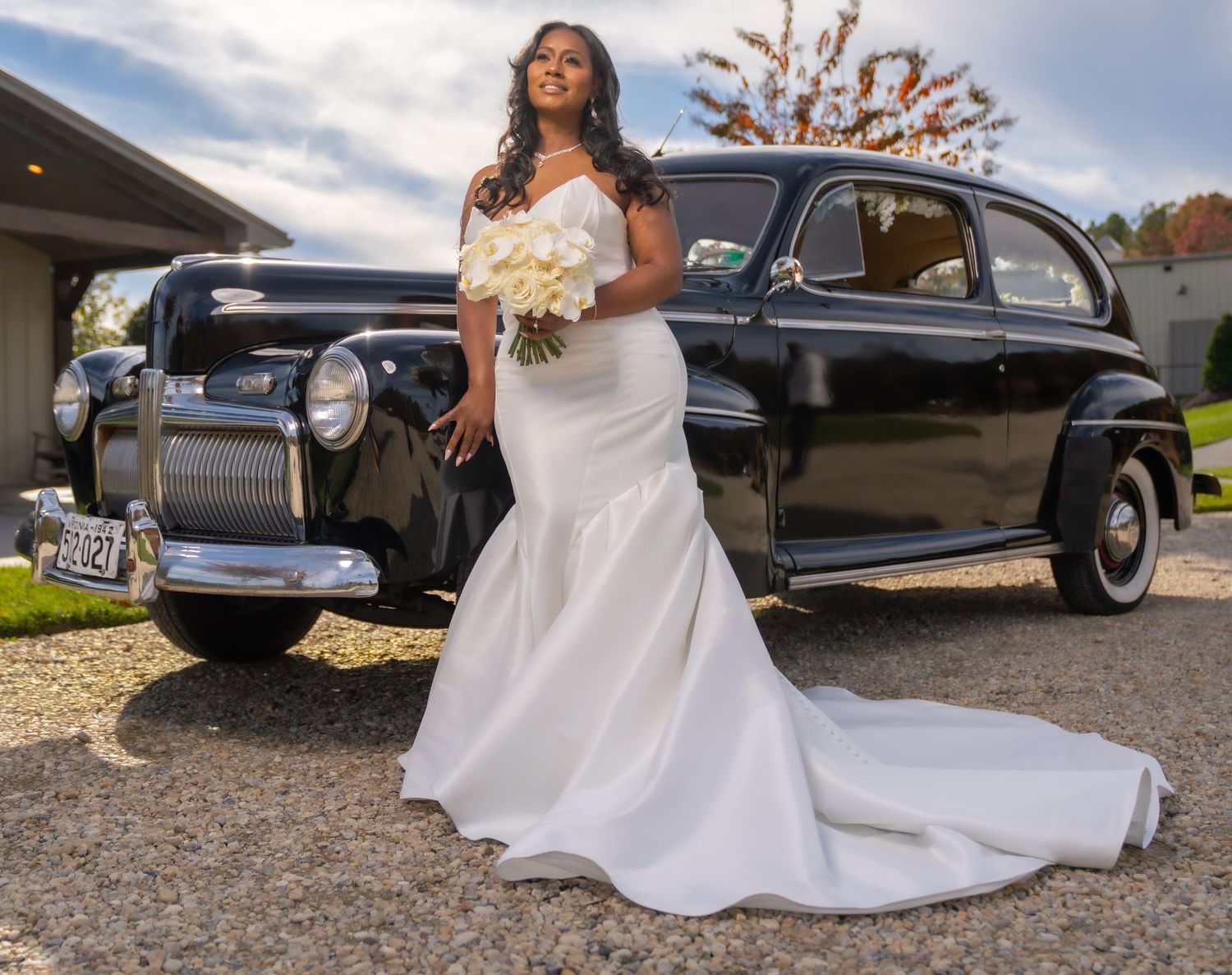 Bride in white mermaid gown holding bouquet poses beside a classic black vintage car on a paved driveway.