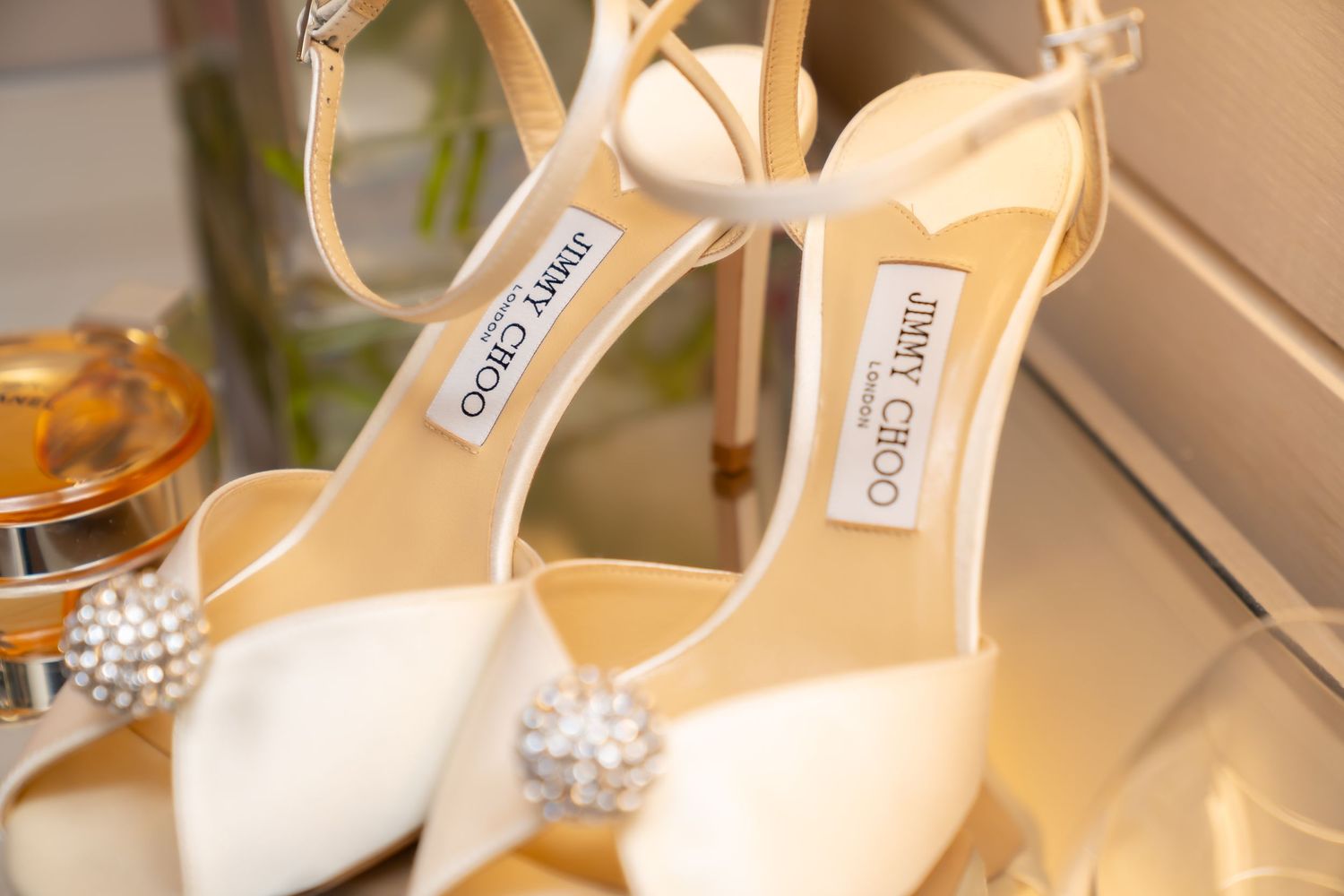 Elegant white Jimmy Choo heels with crystal embellishments and ankle straps displayed on a reflective surface.