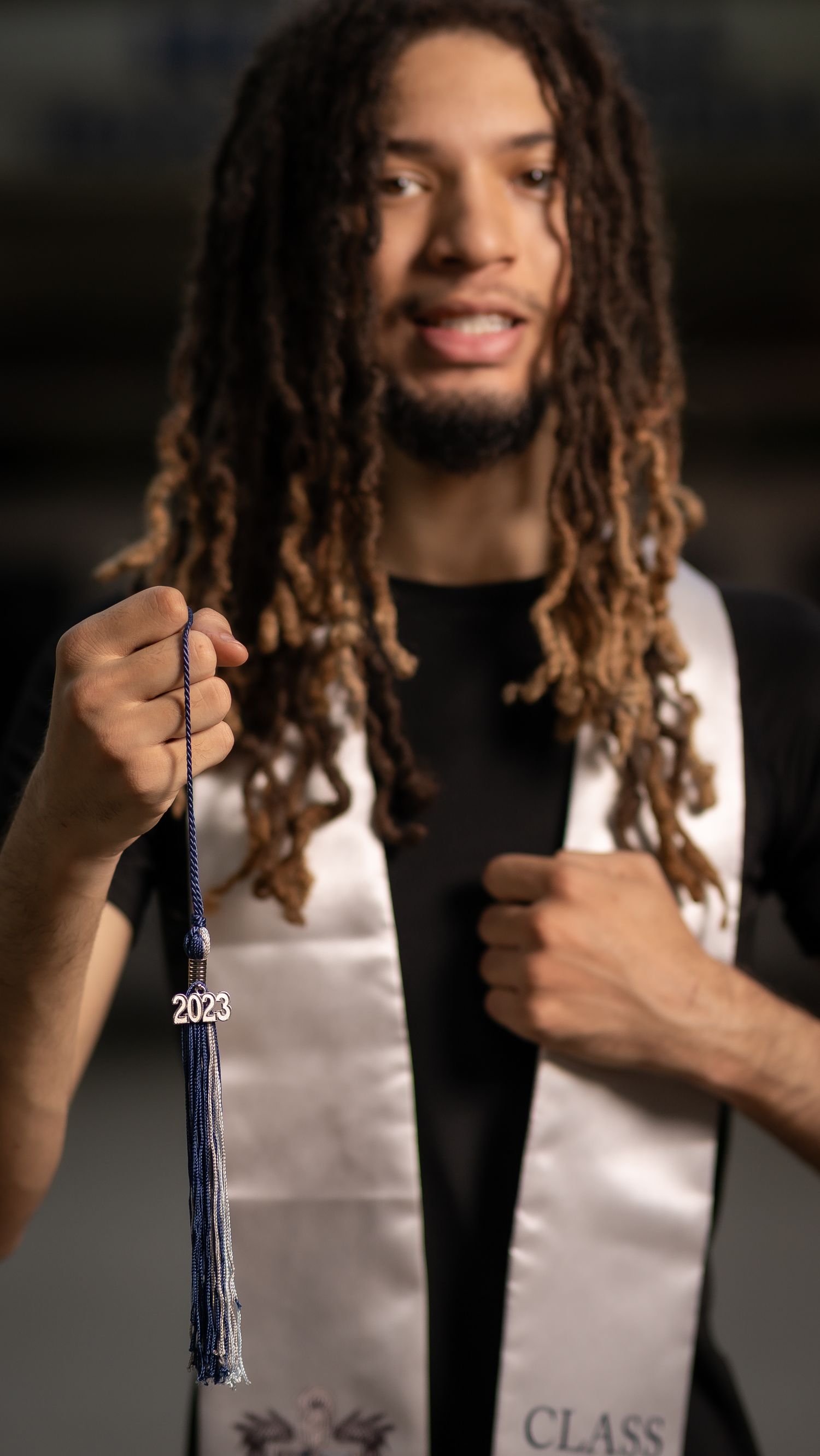 A graduate with long dreadlocks wearing a white graduation stole and holding a 2023 tassel poses for a celebratory photo.