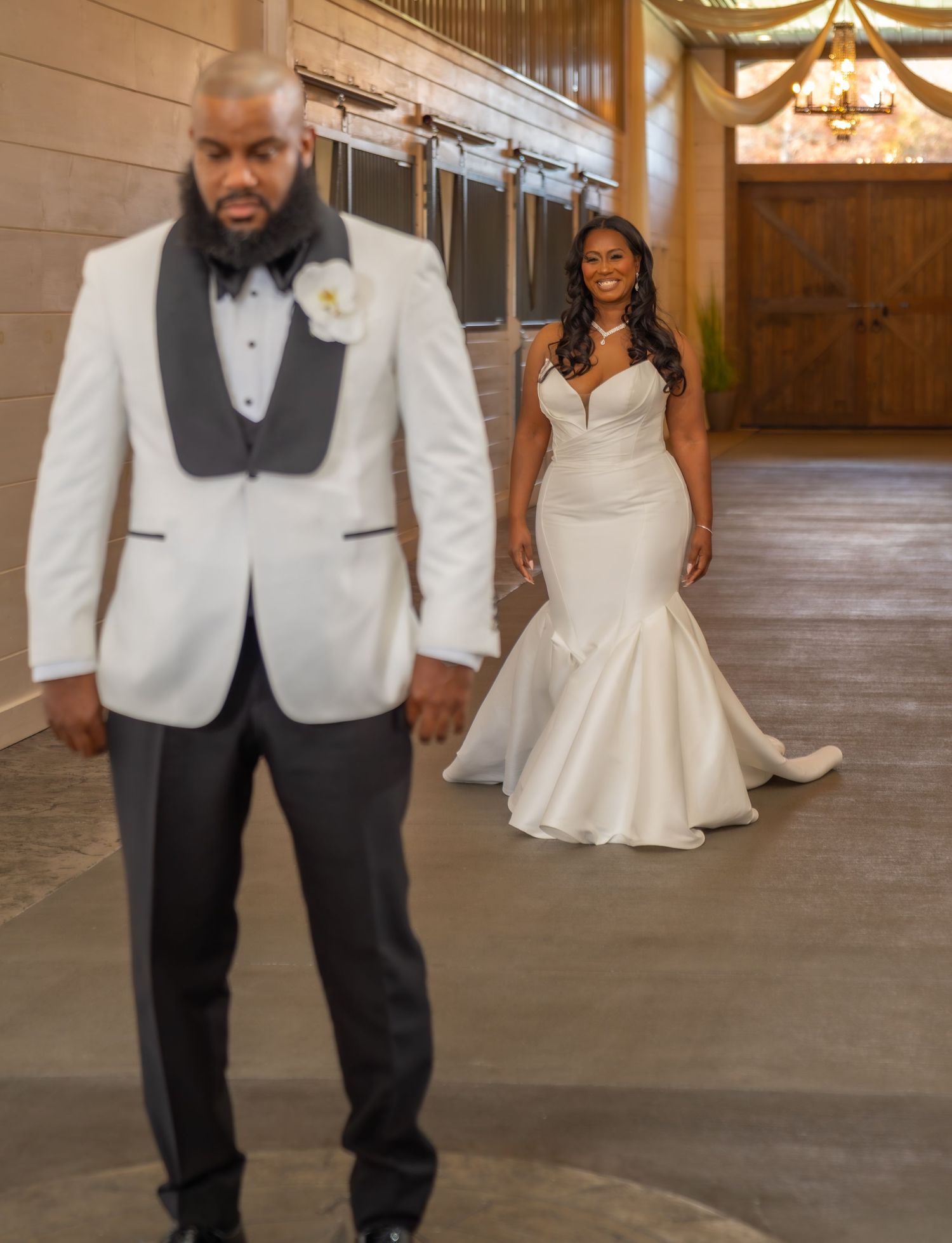 A groom in a white tuxedo stands as his bride in a mermaid wedding gown approaches during their first look.