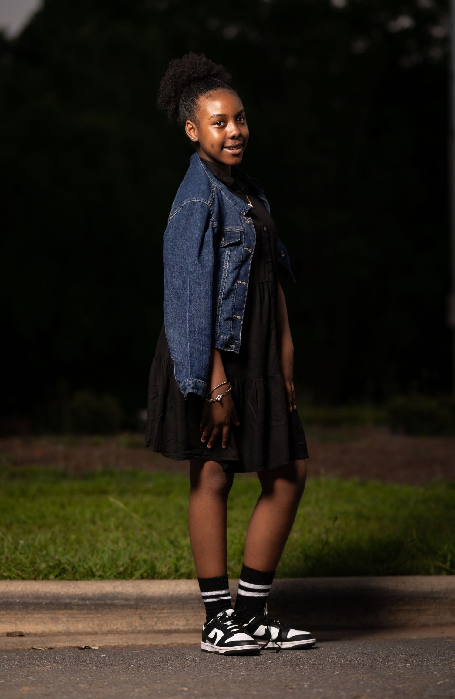 Person wearing a navy denim jacket and black dress poses outdoors with sneakers and crew socks.