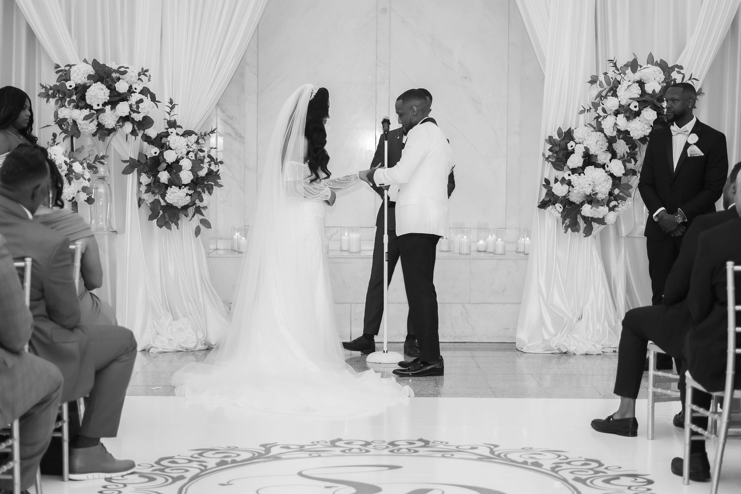 Black and white photo of a couple at the altar during their wedding ceremony surrounded by floral arrangements.