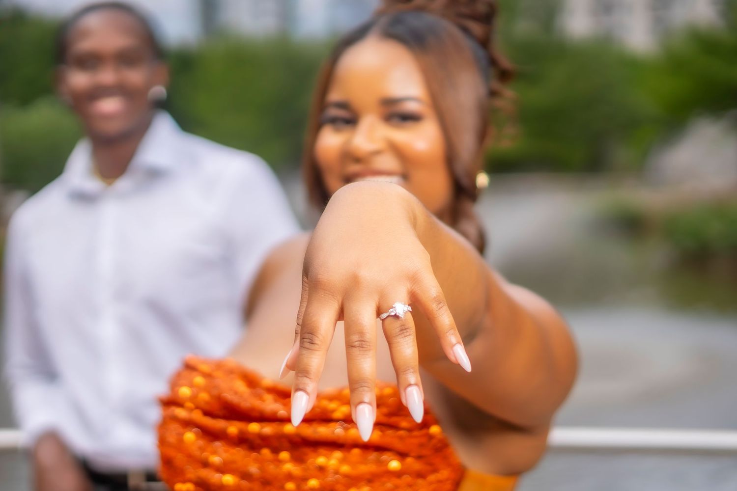 Happy couple celebrating engagement, woman showing off diamond ring with long nails, man smiling in background outdoors.