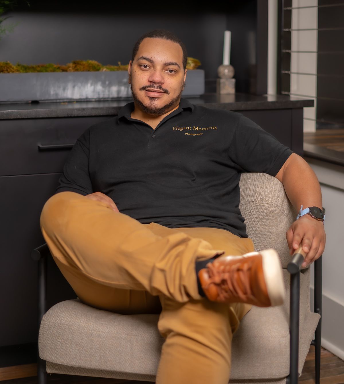 Professional photo of someone seated comfortably in a modern gray armchair wearing black shirt and tan pants.