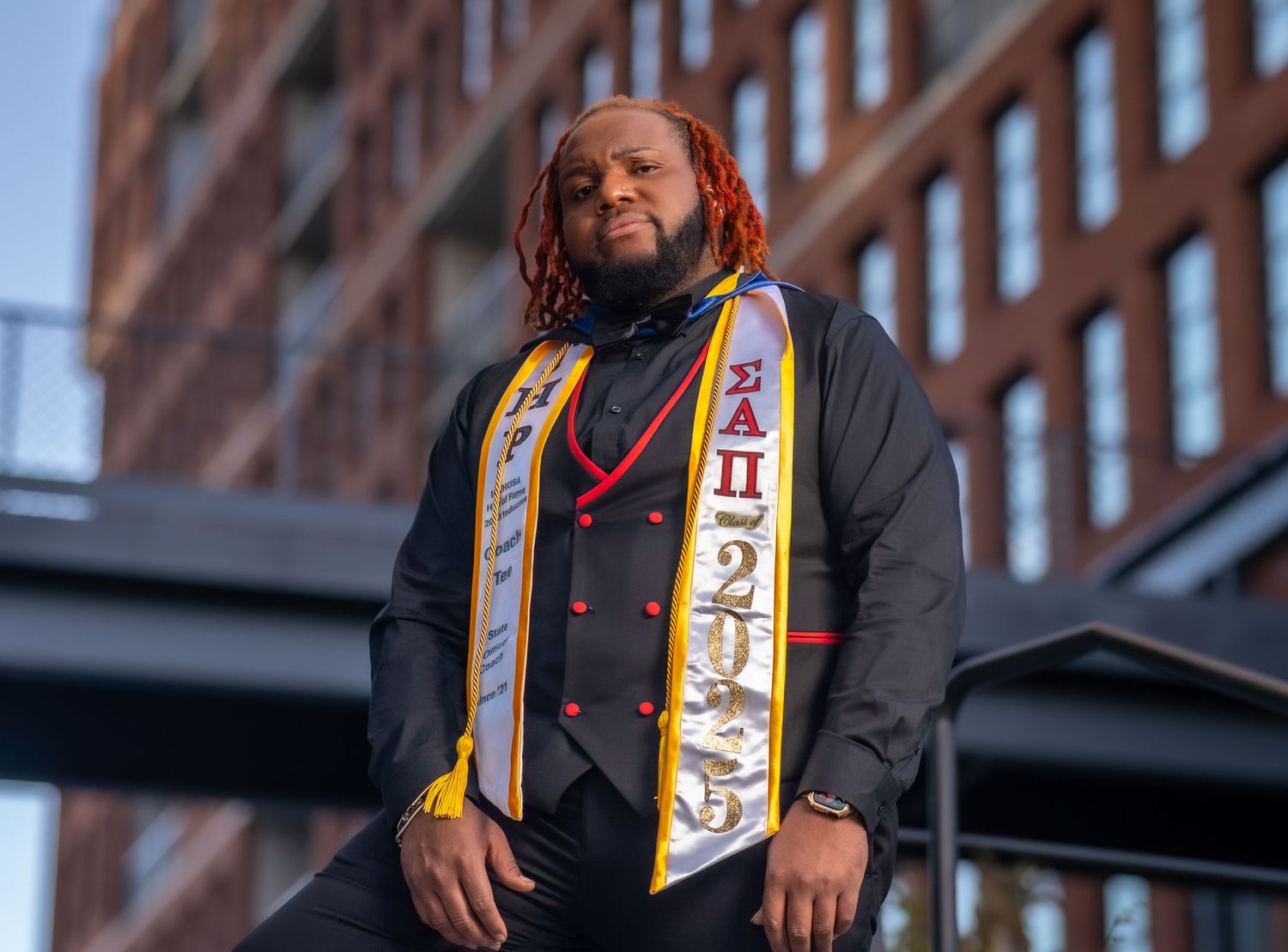 Graduate wearing colorful graduation stole stands in front of brick building at sunset.