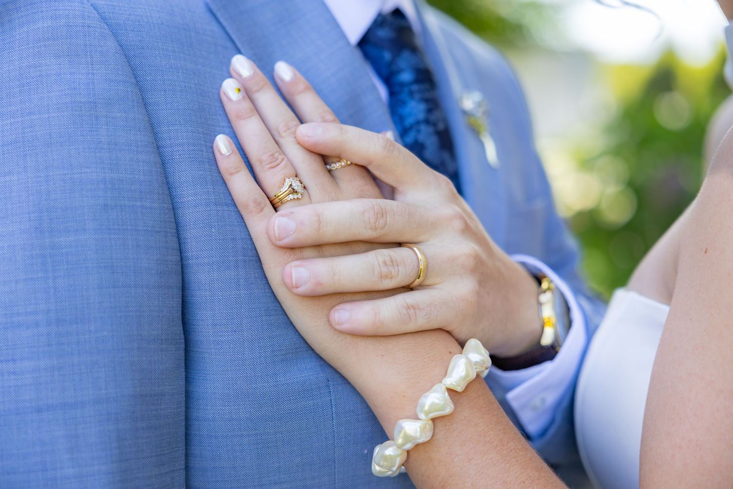 Bride's hand with gold ring and pearl bracelet resting on groom's blue suit jacket at wedding ceremony.
